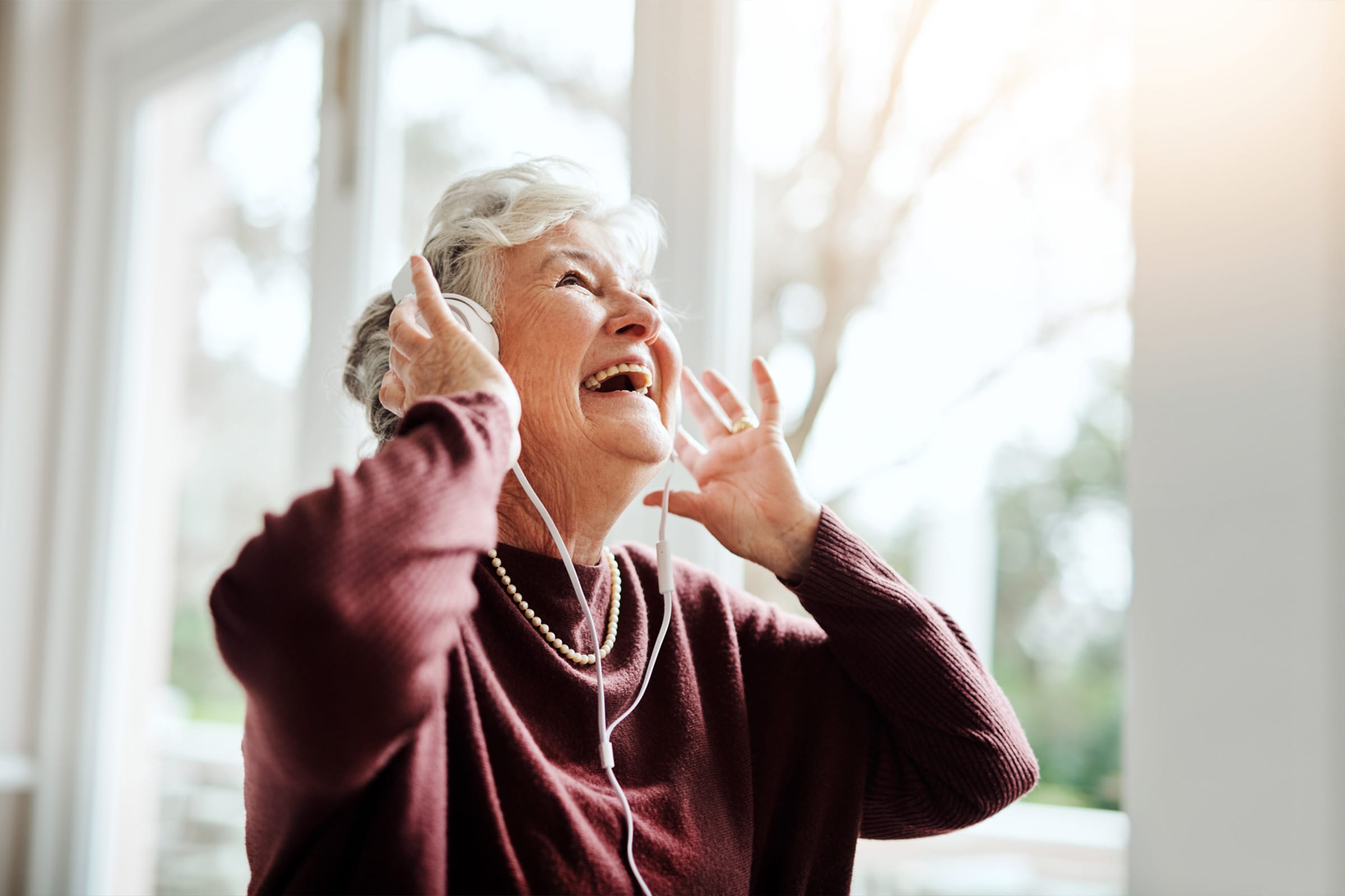 An elderly woman wearing headphones smiles and looks up, appearing joyful as she listens to music. She is indoors, dressed in a maroon sweater and pearl necklace, with sunlight coming through large windows behind her.
