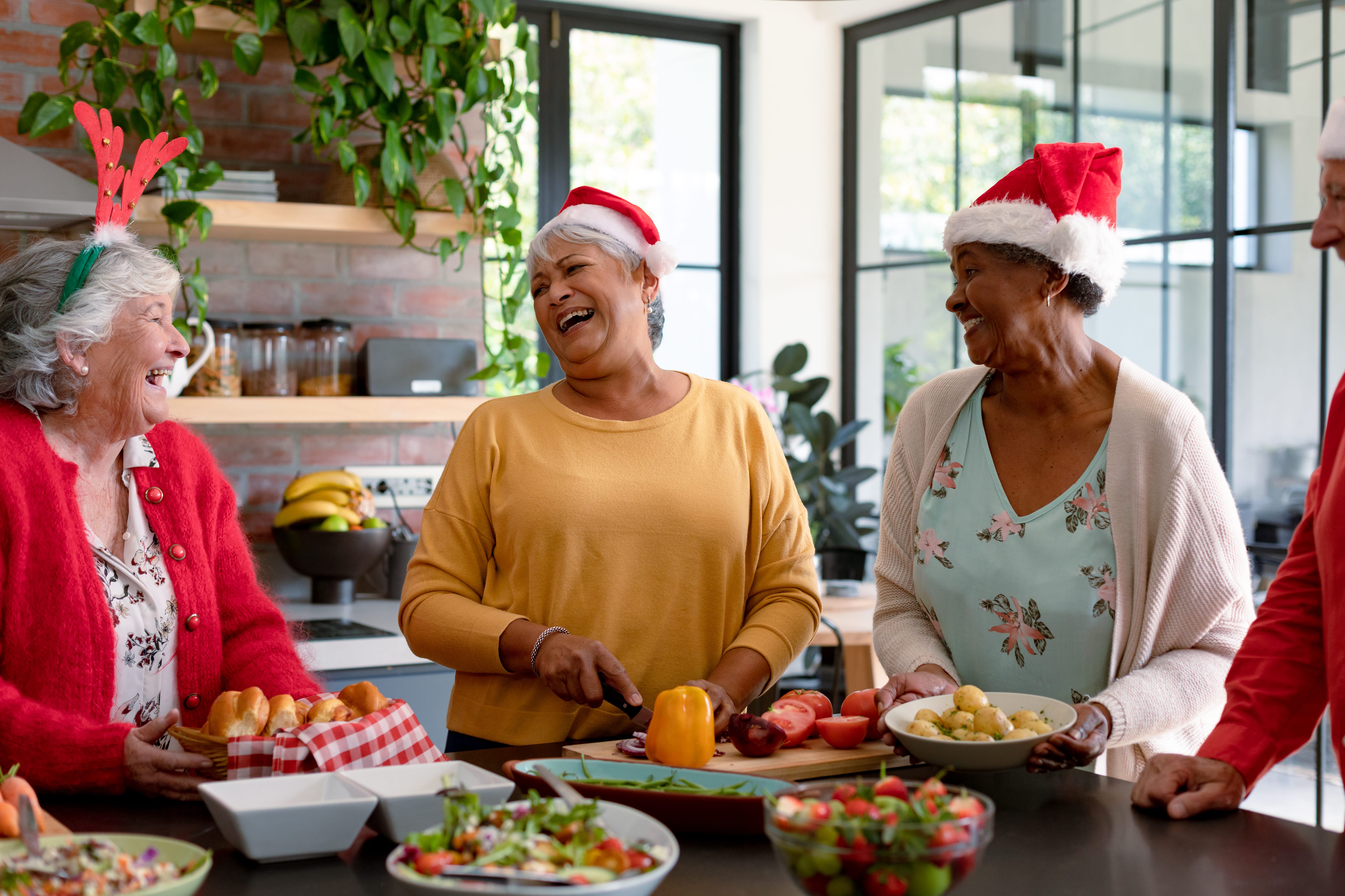 Three older women in festive hats laugh together while preparing food in a bright kitchen. One woman slices a bell pepper, while others hold bowls of bread and potatoes. Holiday decorations are visible in the background.