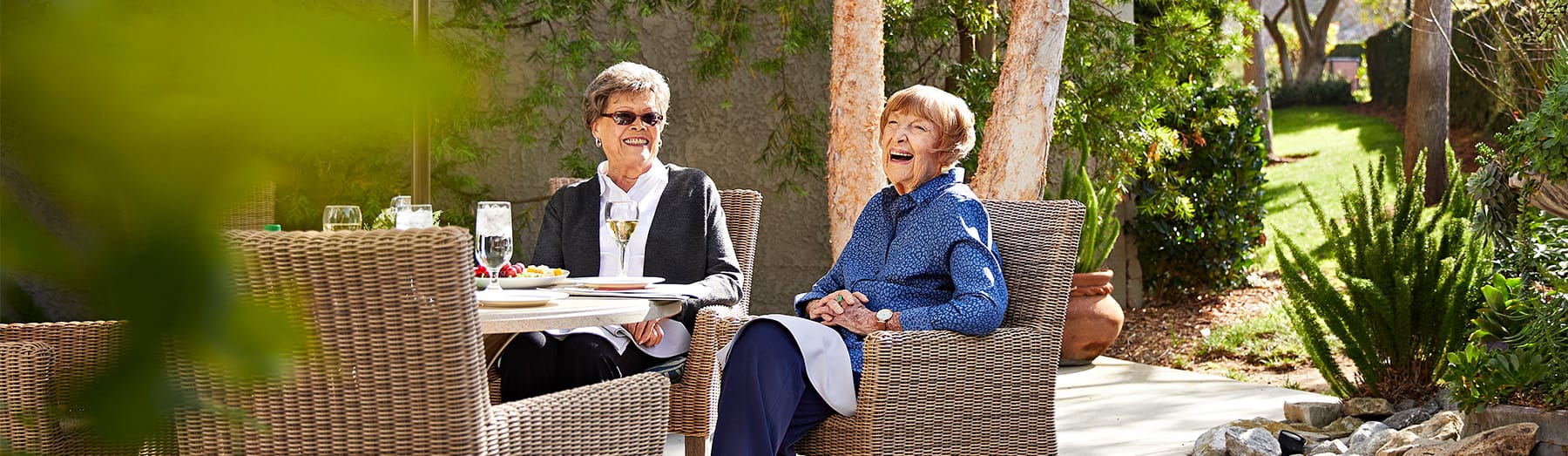 Two older women sit at an outdoor table, smiling and enjoying a meal together in a garden setting with greenery and sunlight around them.
