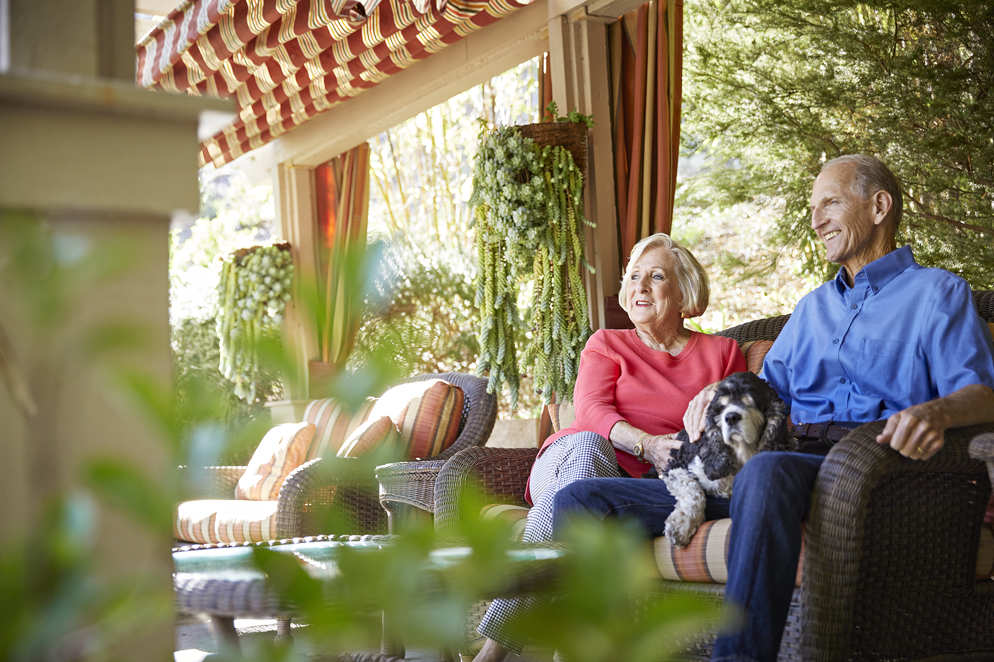An older woman and man sit together on a patio, smiling and relaxing. The woman holds a small black and white dog on her lap. Outdoor furniture and lush greenery surround them. Sunlight filters through the scene.