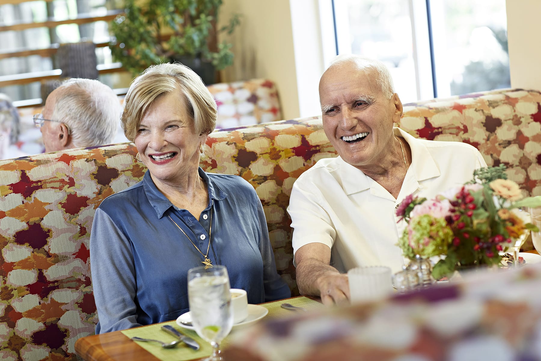 An older man and woman sit together in a colorful restaurant booth, smiling and laughing. A flower arrangement and drinks are on the table in front of them. Other diners are visible in the background.