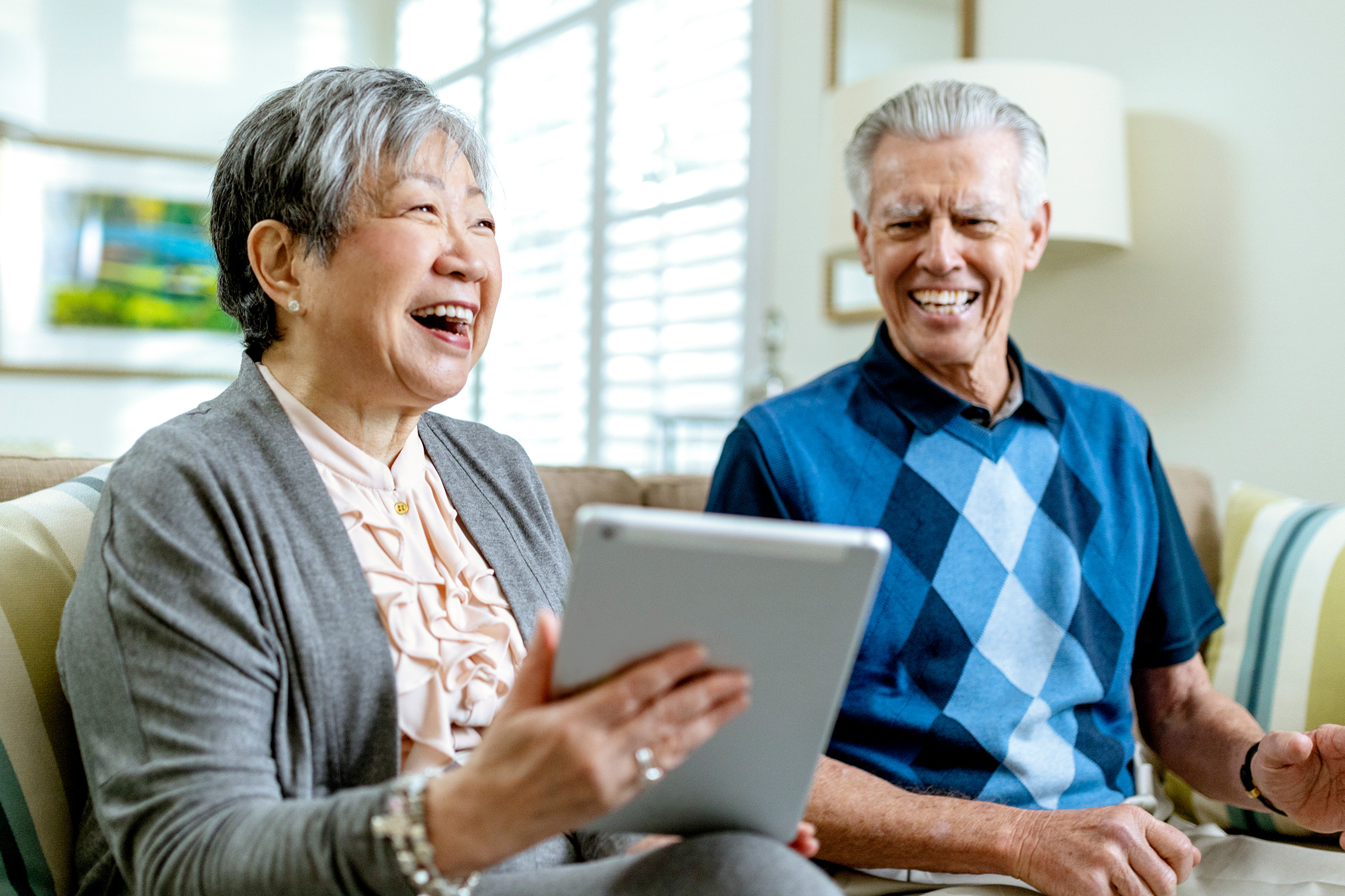 An older woman and man sit on a couch, smiling and laughing together while the woman holds a tablet. Sunlight streams through window blinds in the background, creating a bright, cheerful atmosphere.