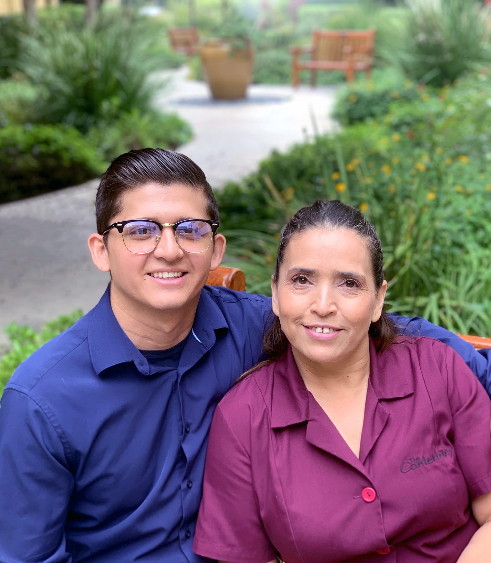A smiling young man in glasses and a navy shirt sits next to a smiling woman in a maroon shirt, with lush greenery and benches in the blurred background of an outdoor setting.