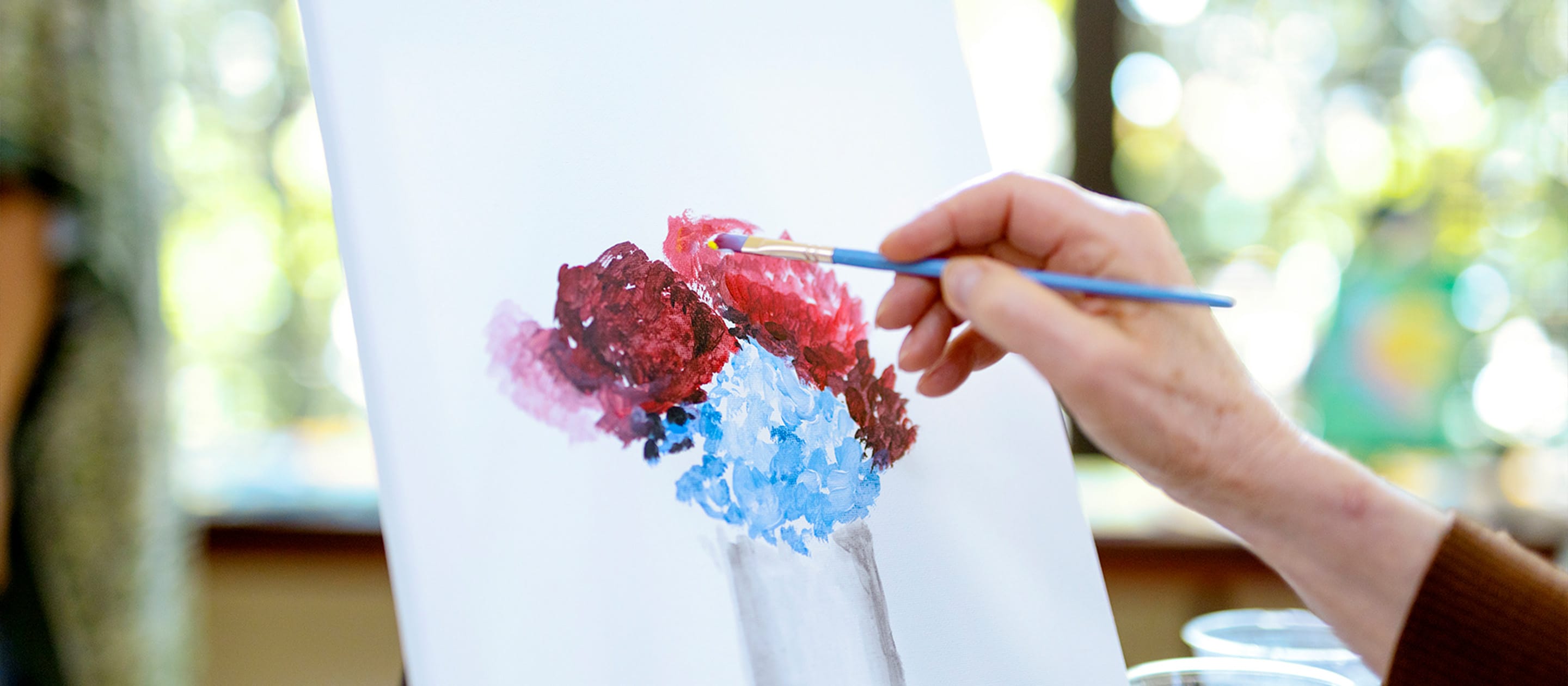 A person’s hand paints a bouquet of red and blue flowers on a white canvas using a paintbrush. The background is softly blurred with natural light and greenery.