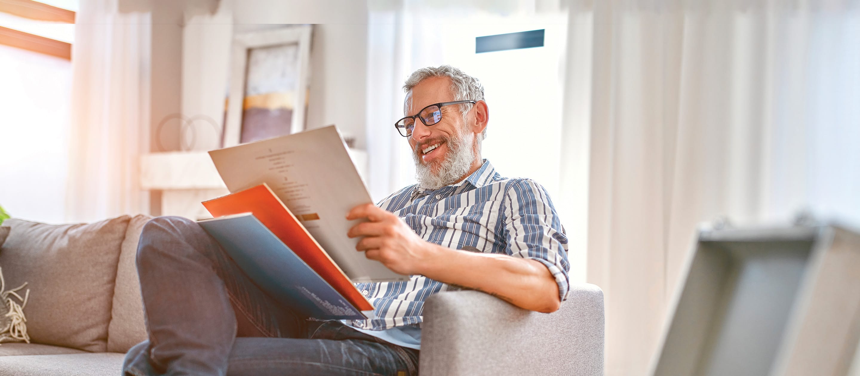 A smiling older man with glasses and a beard sits on a sofa, holding and looking at vinyl records in a bright, cozy living room.