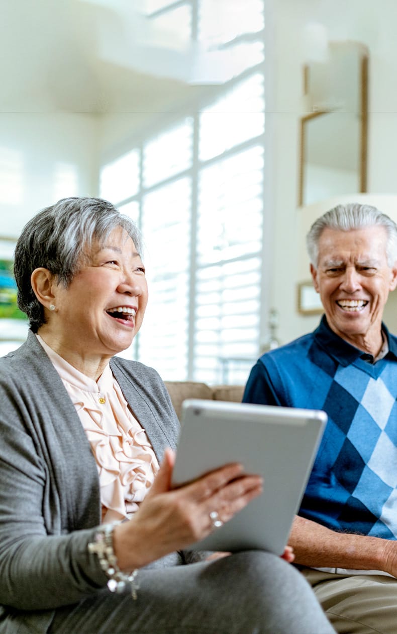 An elderly woman and man sit together indoors, smiling and laughing as the woman holds a tablet. Sunlight streams through large windows behind them, creating a bright and cheerful atmosphere.