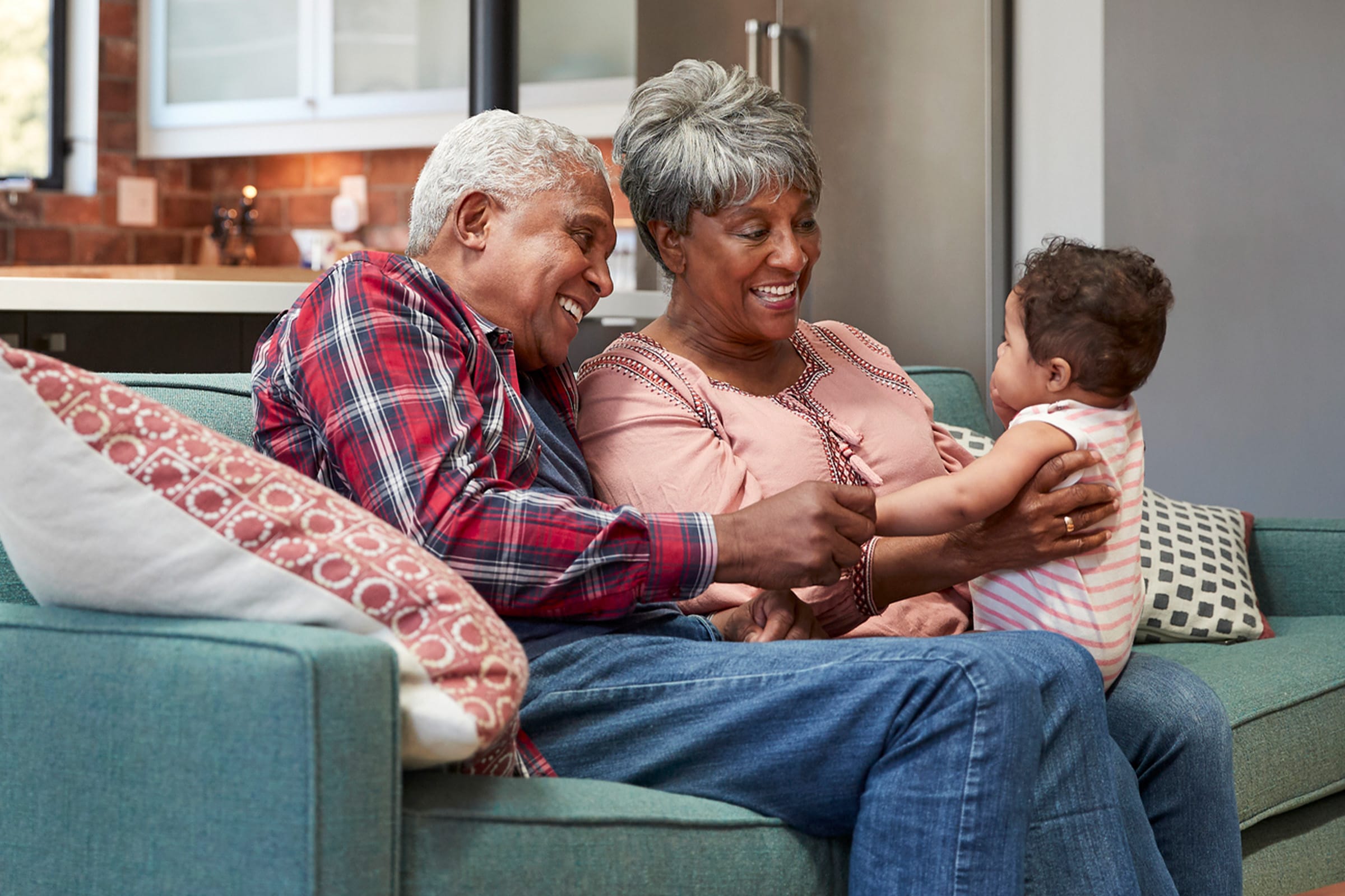 An elderly couple sits on a teal sofa in a cozy living room, smiling and holding hands with a baby who faces them, creating a joyful and warm family moment.