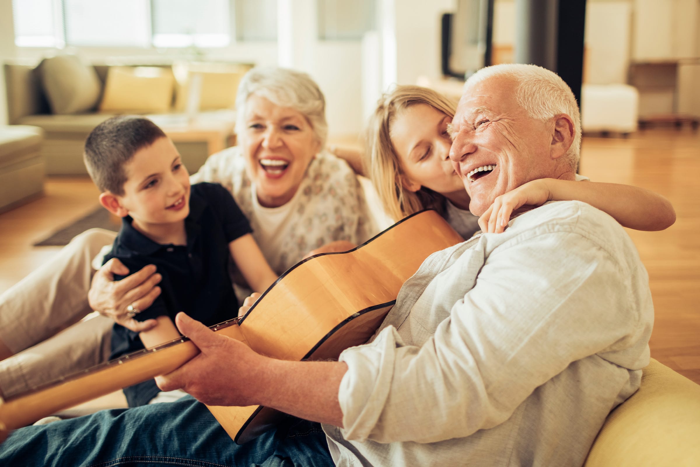 Two grandparents sit on the floor with their two grandchildren, all smiling and laughing. The grandfather plays a guitar while the others gather closely around him in a bright, cozy living room.