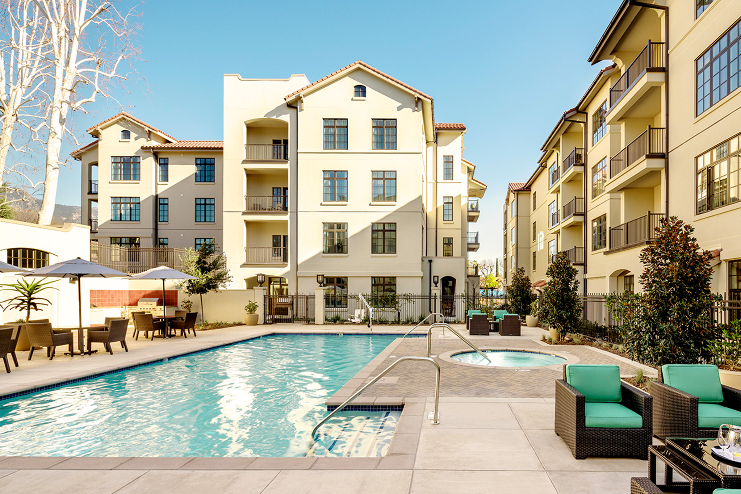 Outdoor swimming pool and hot tub surrounded by lounge chairs, tables, and green cushioned seating, with modern apartment buildings in the background under a clear sky.