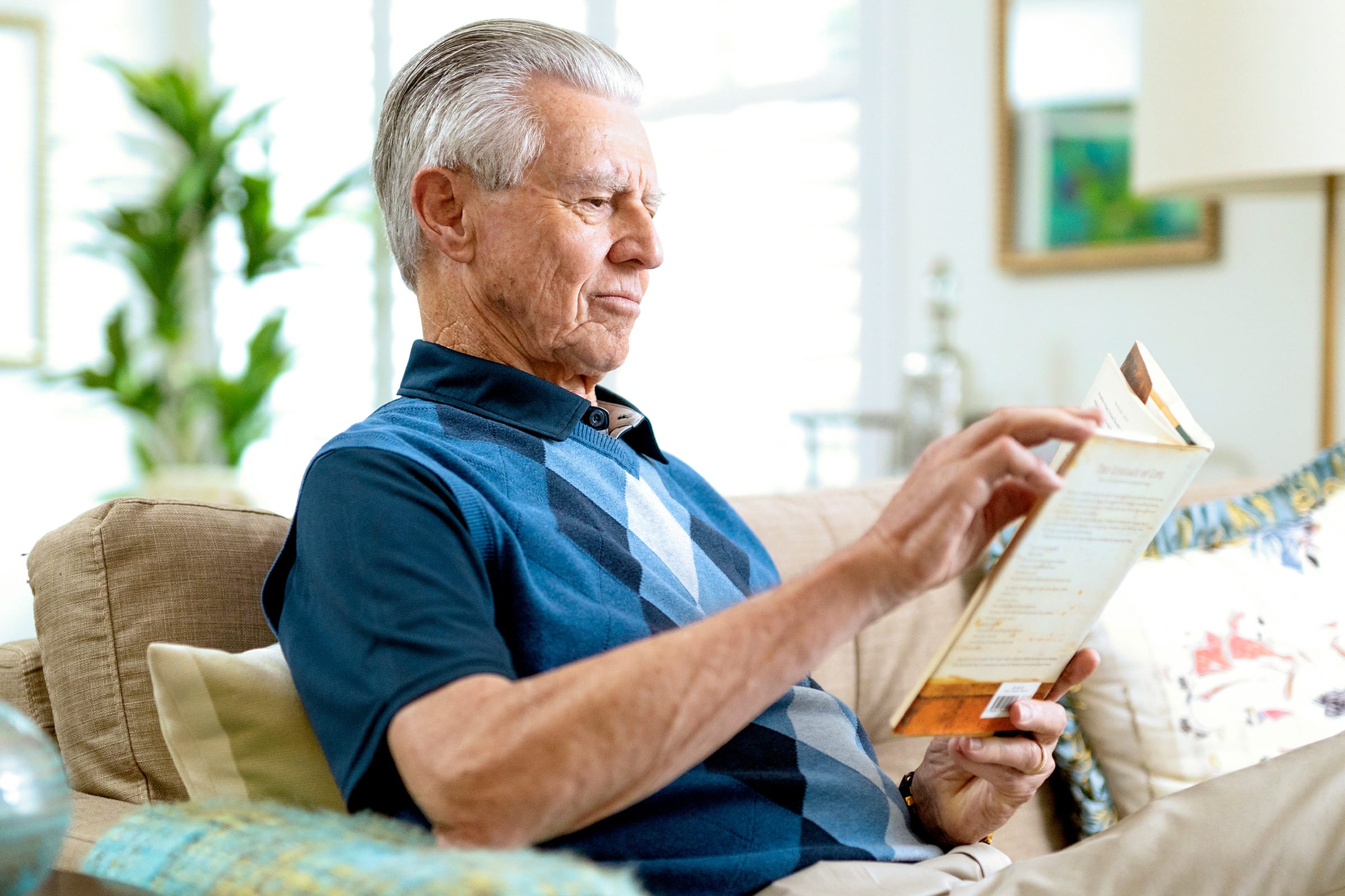 An older man with gray hair sits on a beige couch, wearing a blue argyle polo shirt, reading a book in a brightly lit living room with plants and artwork in the background.