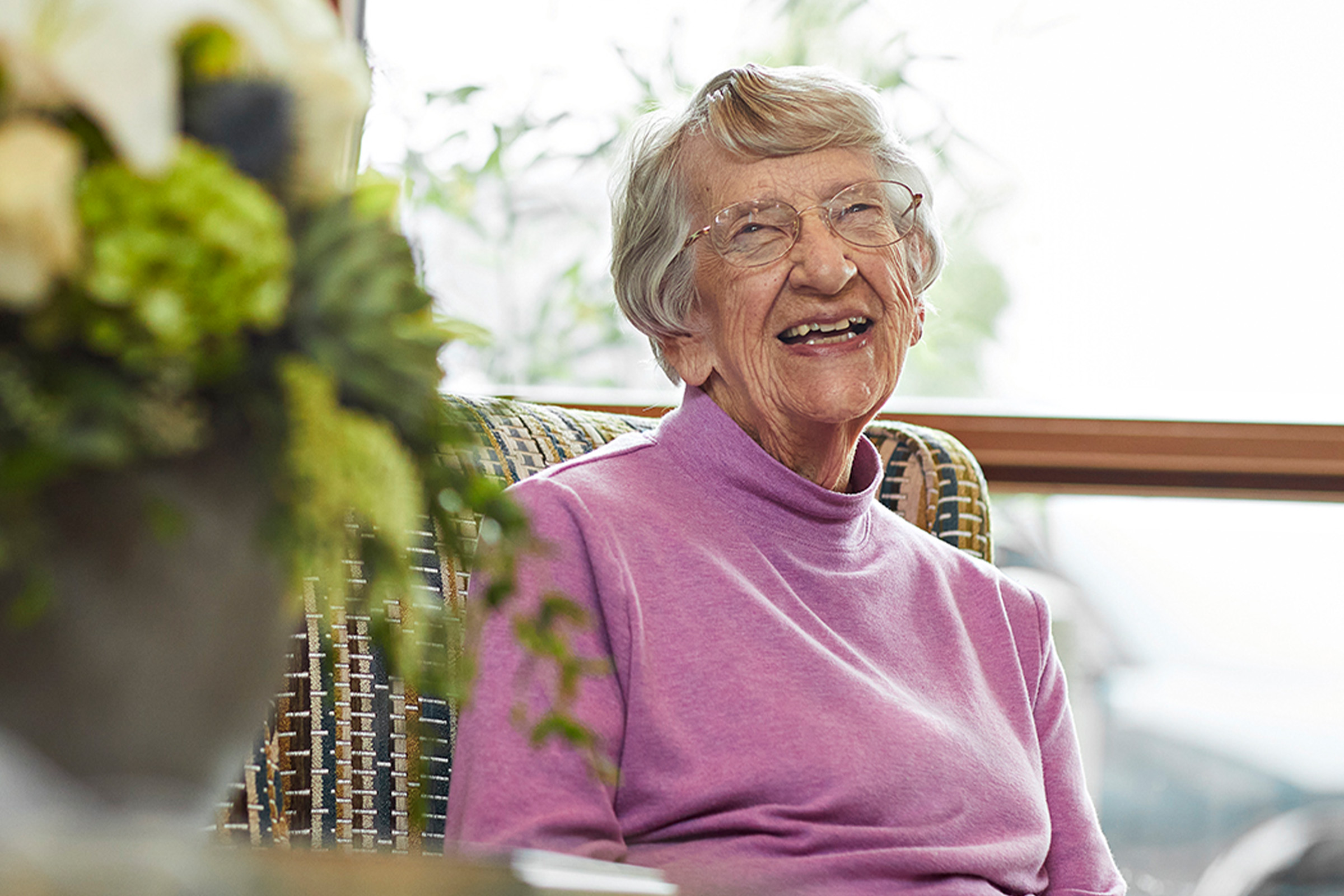 An elderly woman with gray hair and glasses, wearing a light purple turtleneck sweater, sits in a chair and smiles warmly. There is a blurred bouquet of flowers in the foreground.