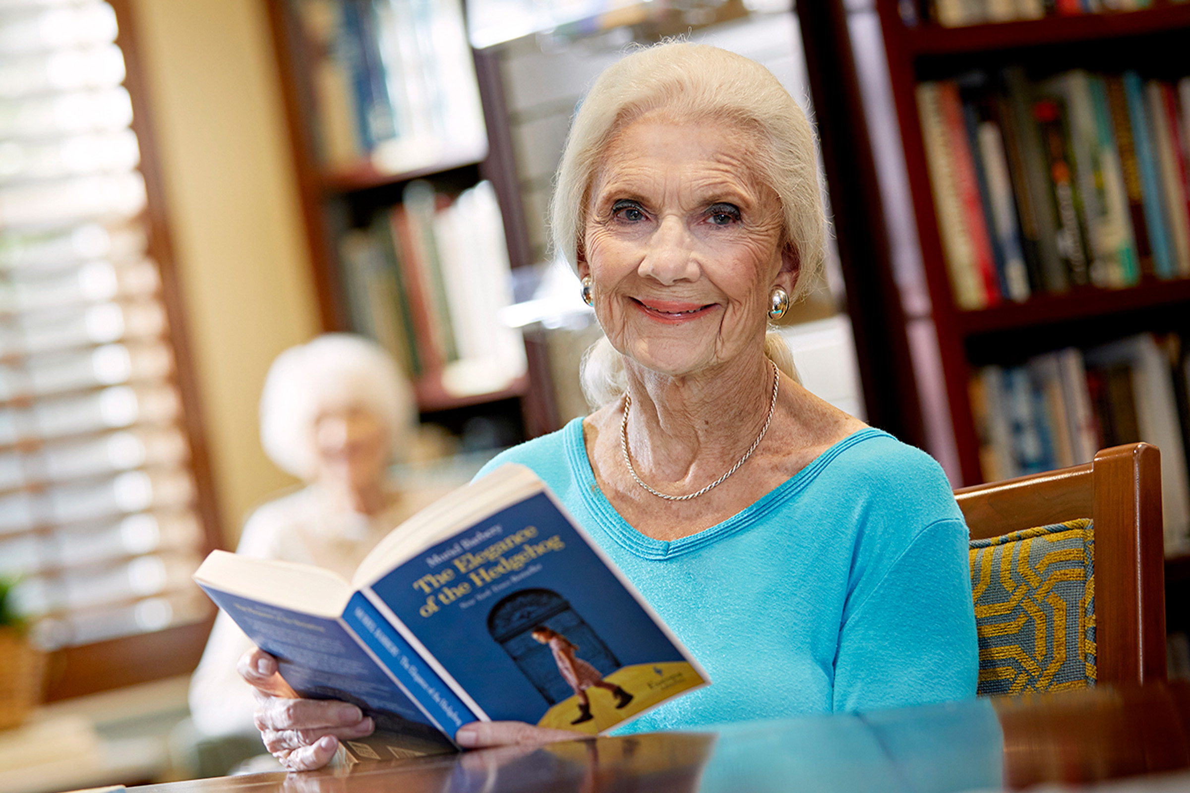 An elderly woman with light hair and a blue top sits at a table, smiling and holding an open book. Bookshelves and another older person are visible in the background.