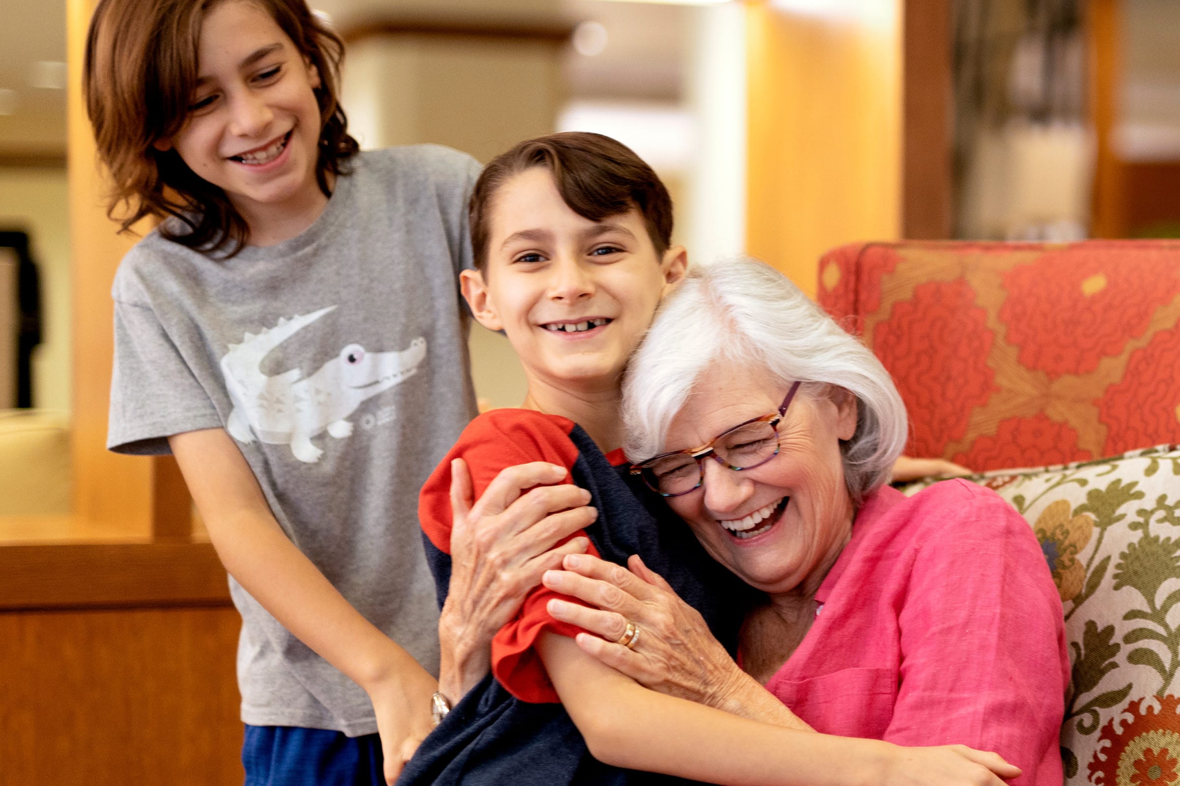 An elderly woman with white hair and glasses sits on a patterned chair, smiling and hugging a young child, while another child stands beside them, all laughing and enjoying a happy moment together indoors.
