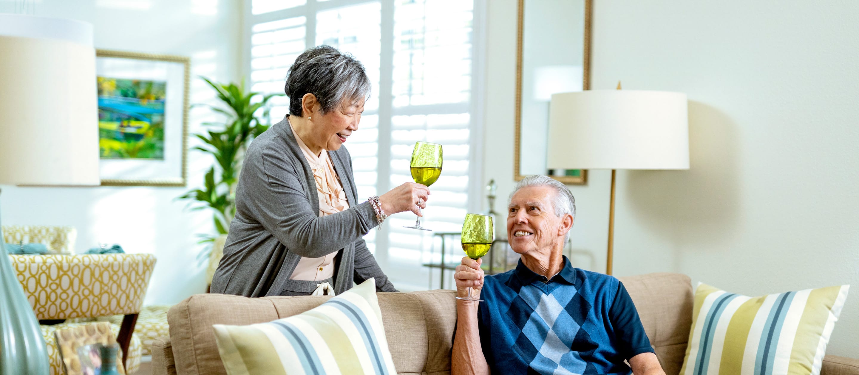 An older woman and man smile and toast with green wine glasses while sitting on a beige couch in a bright, cozy living room with striped pillows and sunlight streaming through the windows.