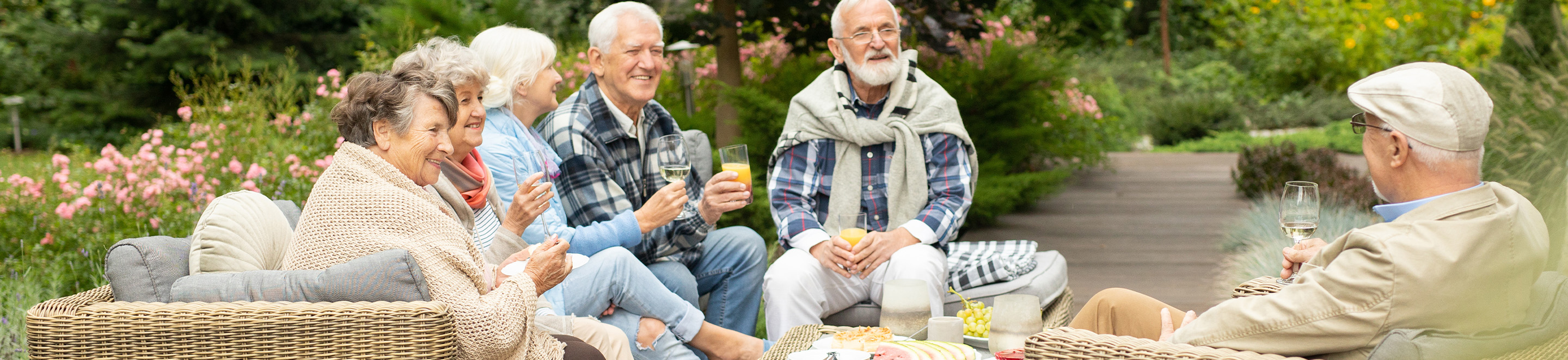A group of six smiling older adults sit outdoors on wicker furniture, chatting and holding drinks. They are surrounded by lush greenery and pink flowers, enjoying a relaxed and friendly gathering.