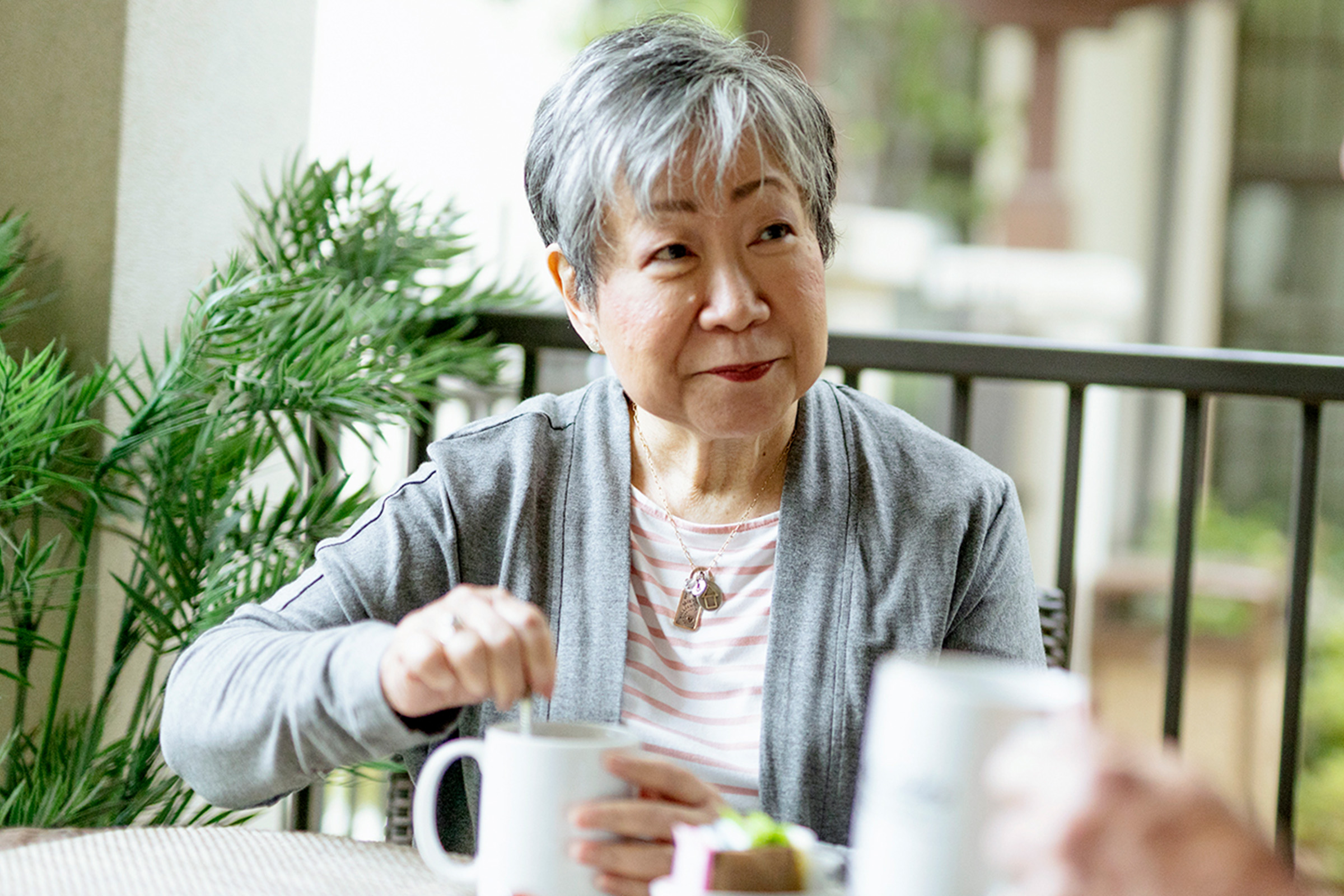 An older woman with short gray hair sits at a table outdoors, stirring a drink in a white mug. She is wearing a gray cardigan over a striped shirt and is smiling while talking to someone off-camera. Plants are in the background.