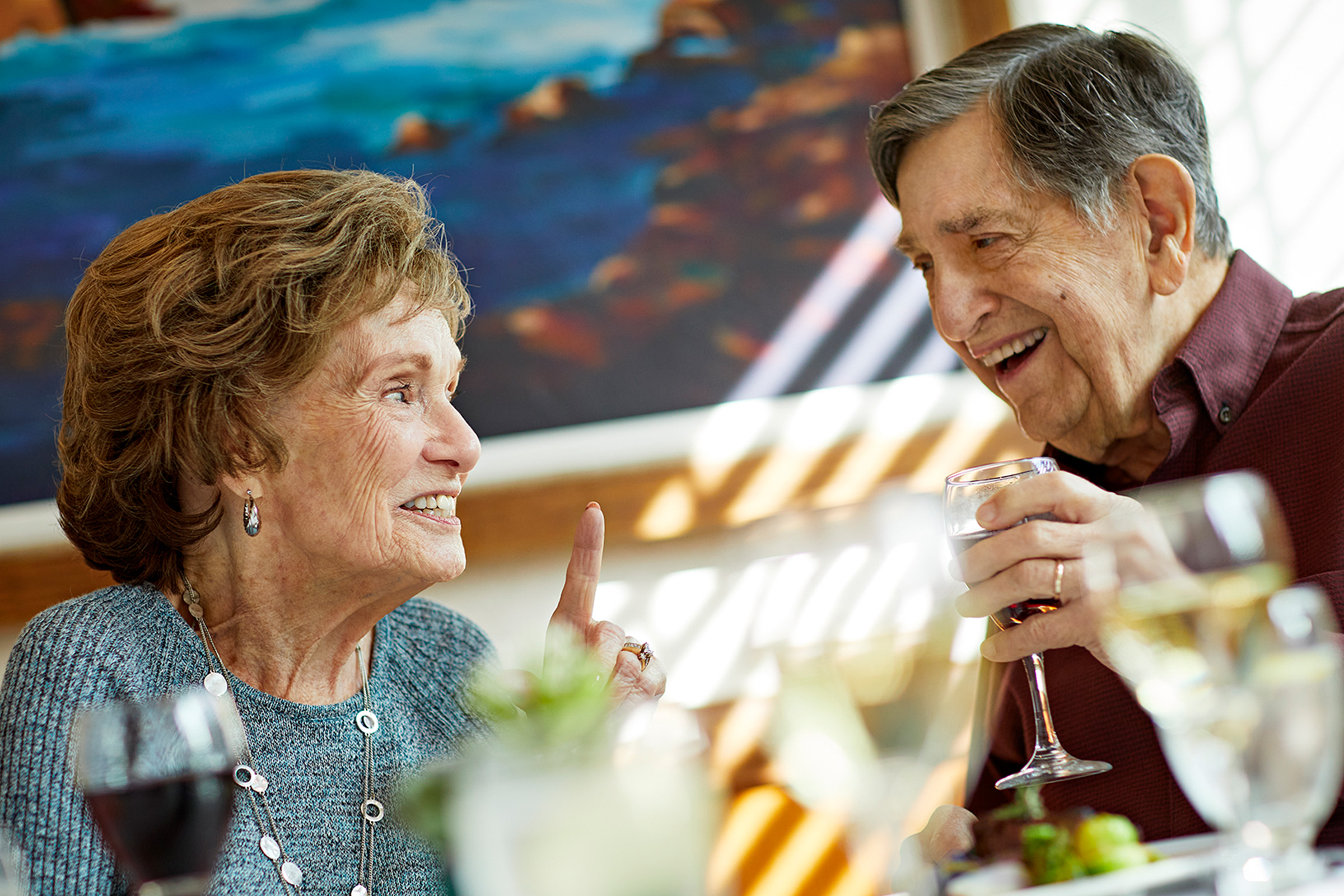 An elderly woman and man sit together at a table, smiling and talking. The woman gestures with her finger raised, while the man holds a glass of wine. They appear to be enjoying a cheerful conversation.