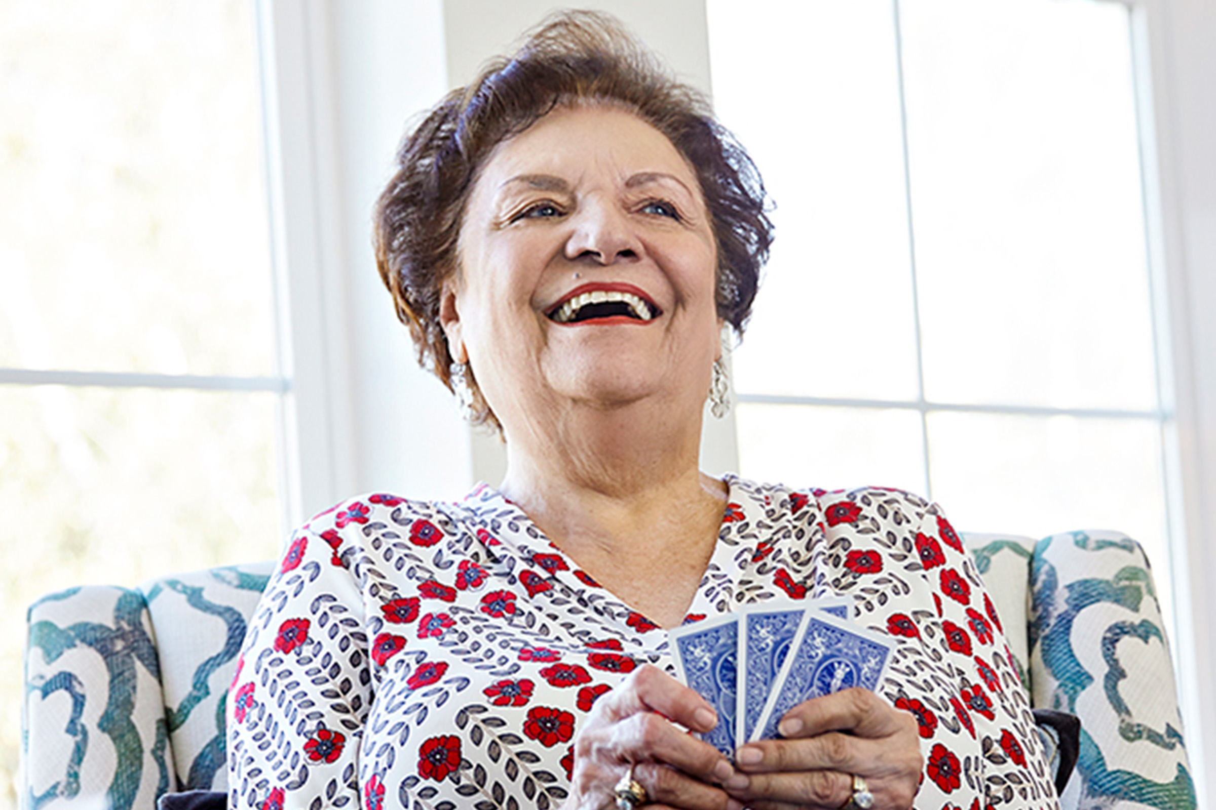 An older woman with short brown hair smiles and laughs while holding playing cards, sitting indoors on a patterned chair in front of a bright window.