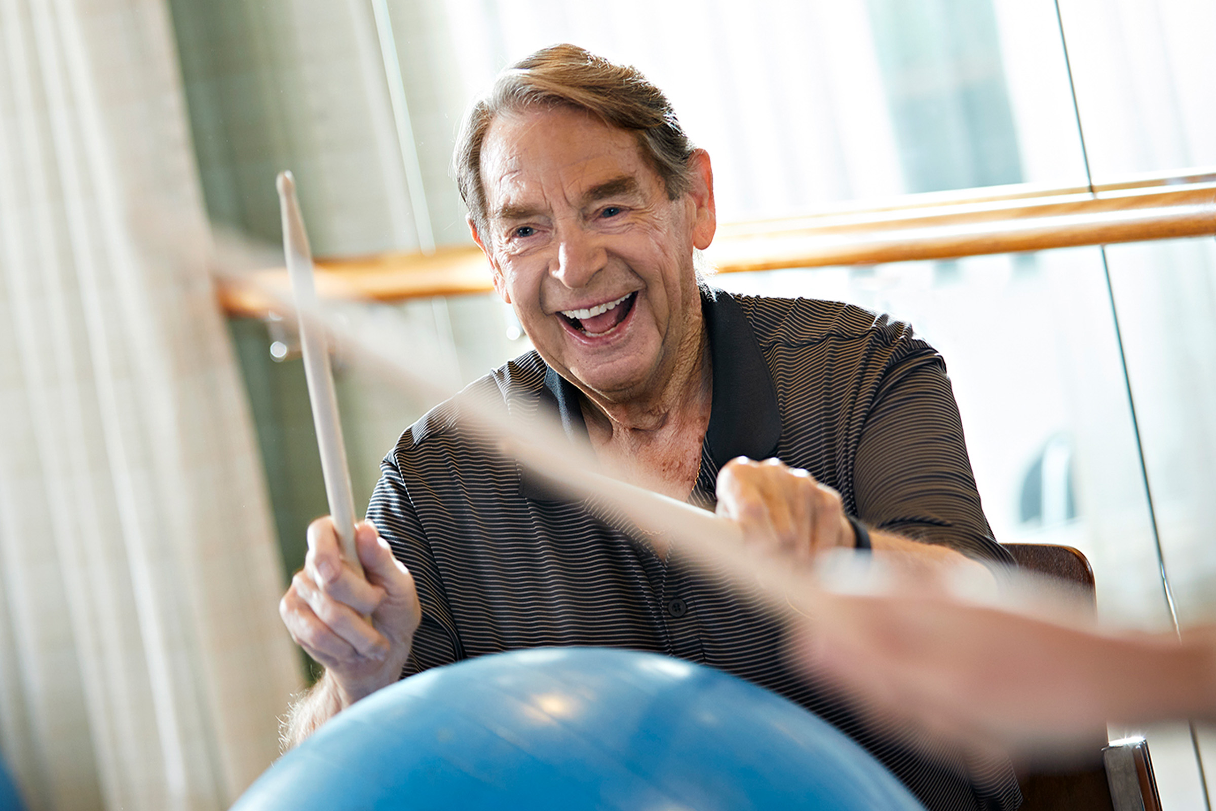 Demonstrating independent living in Orange County, a smiling resident uses drumsticks on a exercise ball.