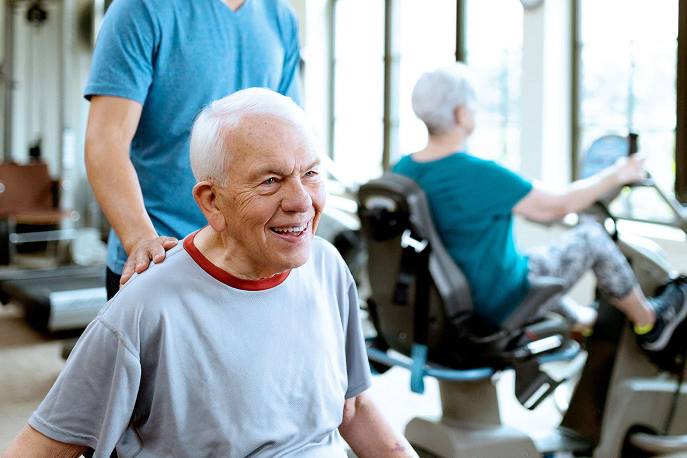 An elderly man smiles while being assisted by a caregiver. In the background, an elderly woman exercises on a stationary bike in a bright, sunlit gym or rehabilitation center.