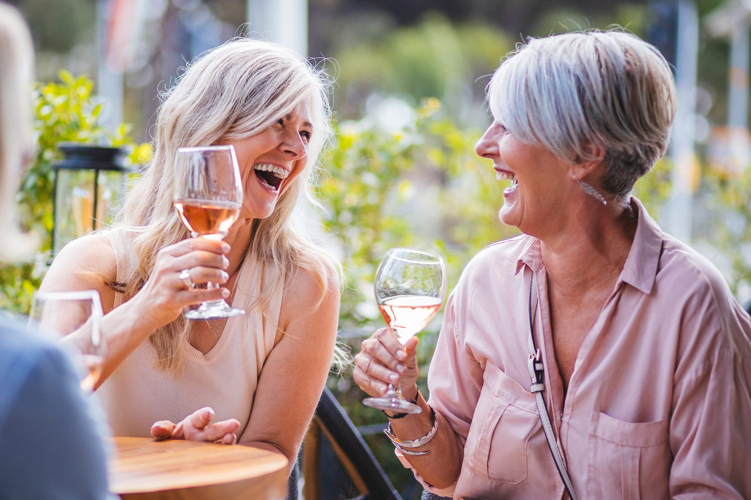 Two women sit outdoors, smiling and laughing while holding glasses of rosé wine. They appear to be enjoying a lively conversation on a sunny day, surrounded by greenery.