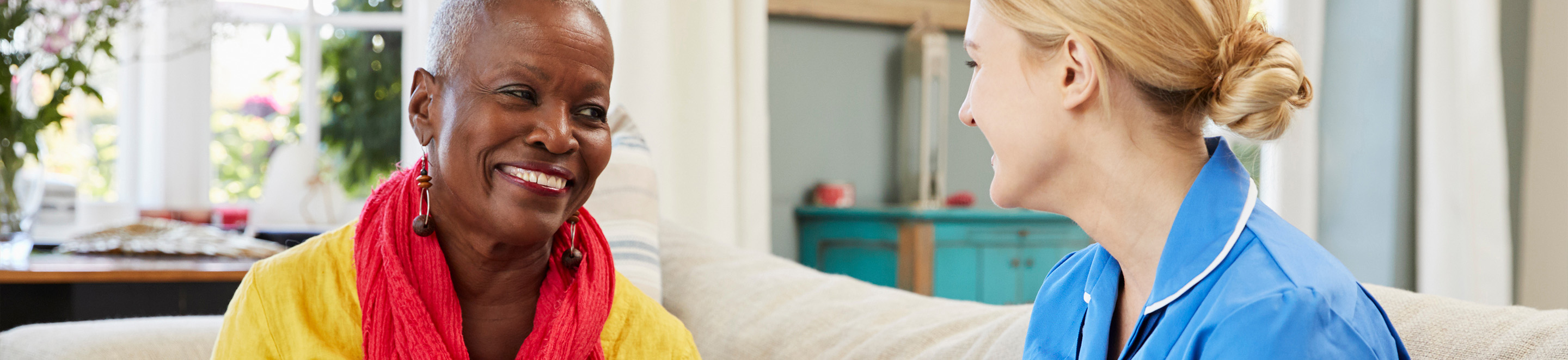 An elderly woman wearing a yellow top and red scarf smiles while talking with a caregiver in a blue uniform sitting beside her on a couch in a cozy living room.