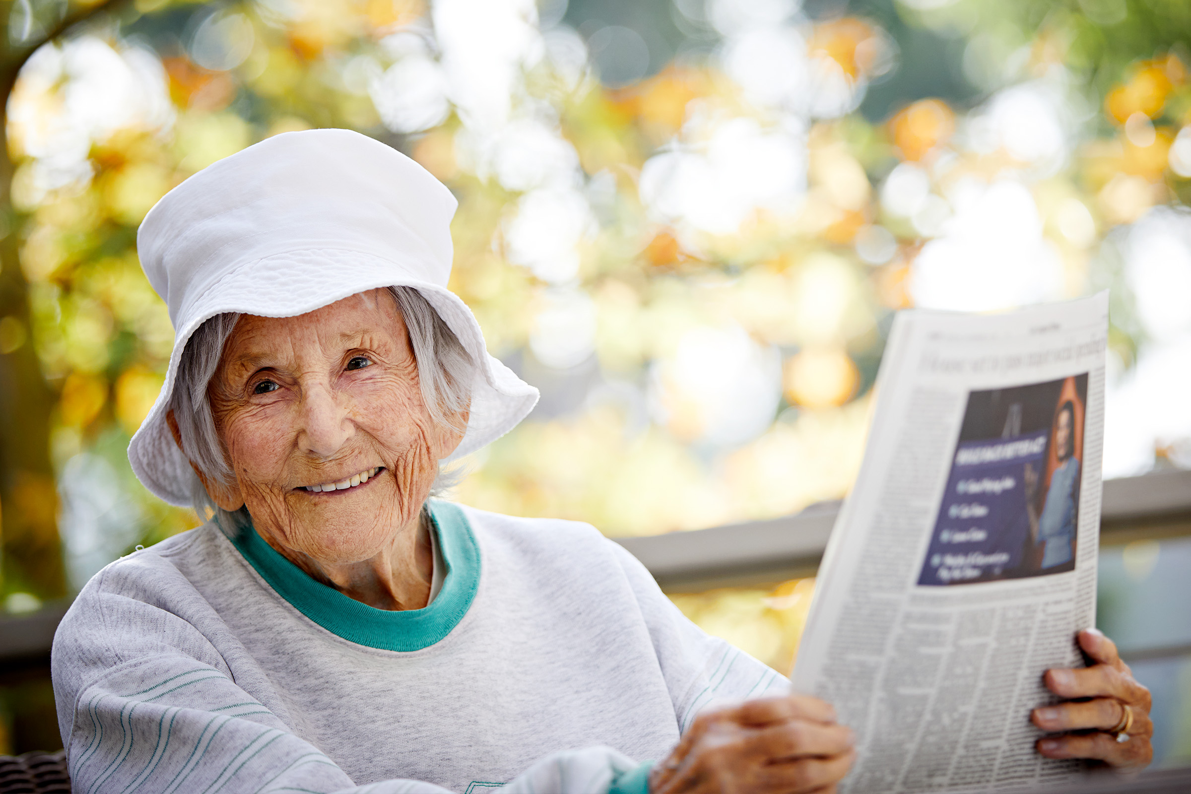 1.5_CAN_Financial An elderly woman wearing a white sun hat and a gray sweatshirt smiles while sitting outdoors and reading a newspaper. Trees with green and yellow leaves are blurred in the background.