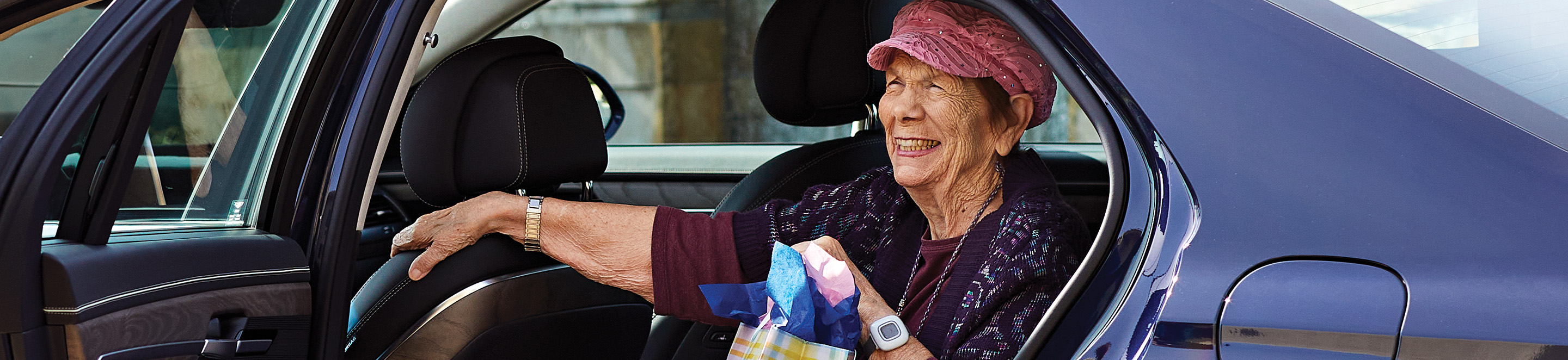 1.5_CAN_hero An elderly woman wearing a pink cap and patterned sweater smiles as she sits in the back seat of a car, reaching out with one hand and holding a colorful bag in the other.