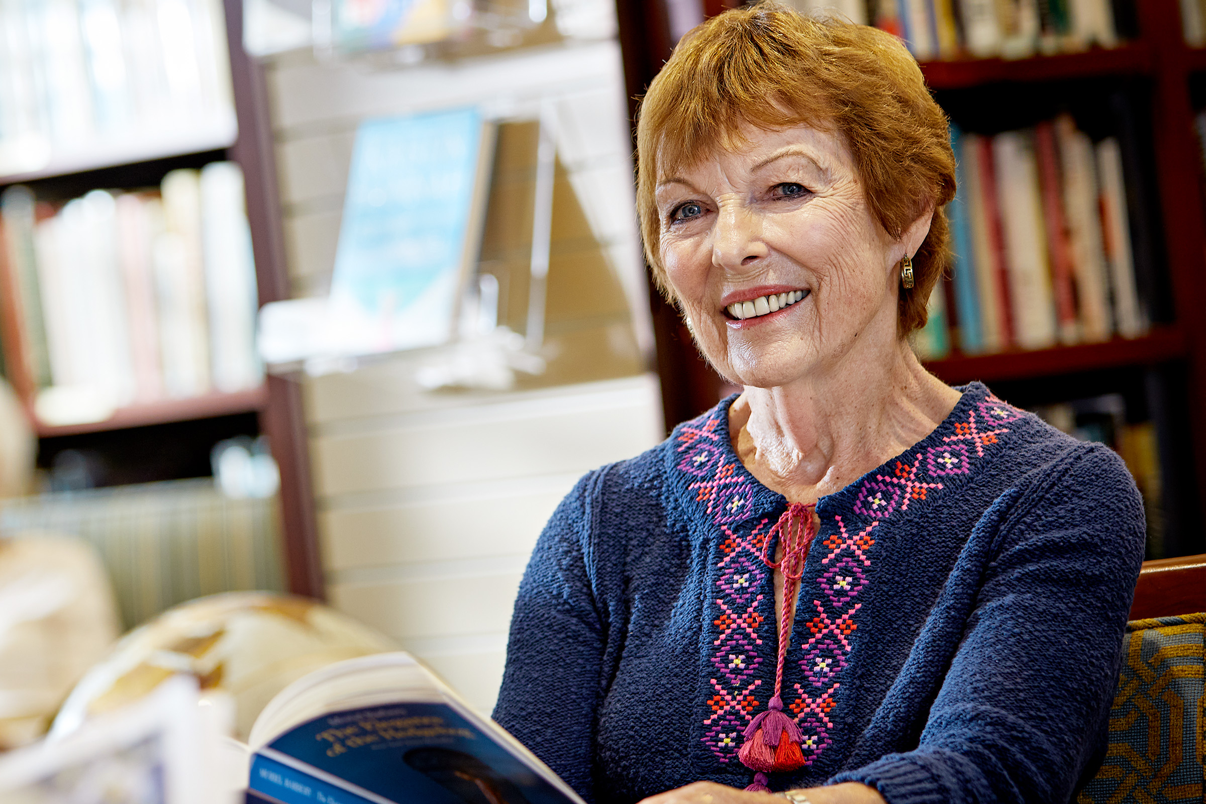 An older woman with short red hair and a blue embroidered top smiles while holding an open book, seated in a cozy room with bookshelves in the background.