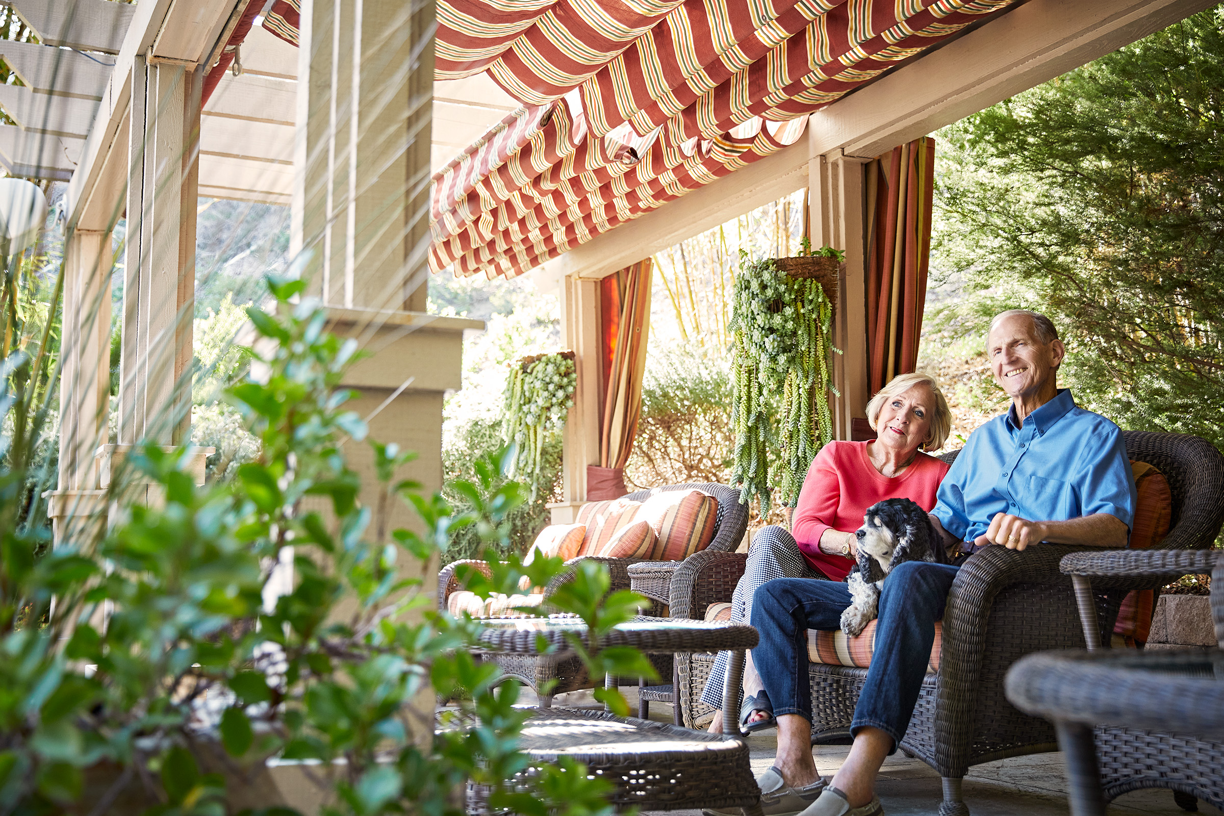 An older couple sits together on a shaded patio, smiling and holding a small dog on their laps. They are surrounded by outdoor furniture, hanging plants, and greenery under a striped awning.