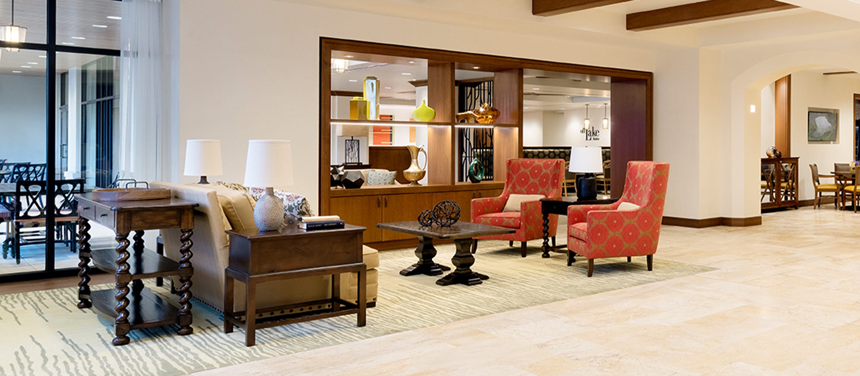 A bright, modern hotel lobby with two red patterned armchairs, a sofa, wood tables, decorative shelves, and artwork. Large windows and light tile floors create an inviting, spacious atmosphere.