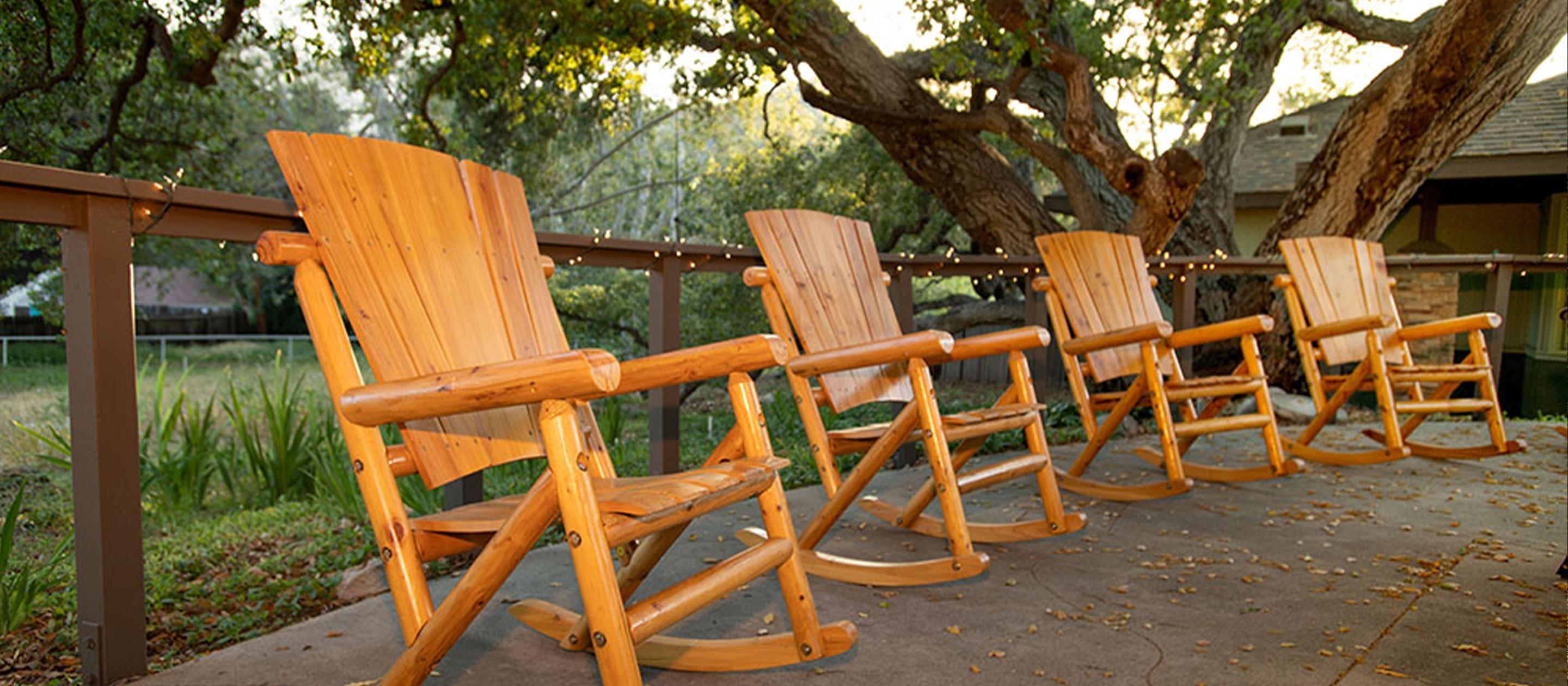 Four wooden rocking chairs are lined up on a shaded patio, overlooking a grassy yard with trees. String lights are draped along the railing, and sunlight filters through the branches.