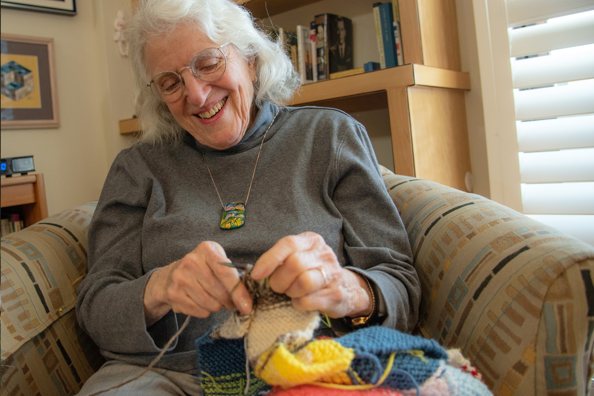 An elderly woman with glasses and white hair smiles while knitting a colorful piece, sitting on a patterned armchair in a cozy room with bookshelves and window blinds in the background.