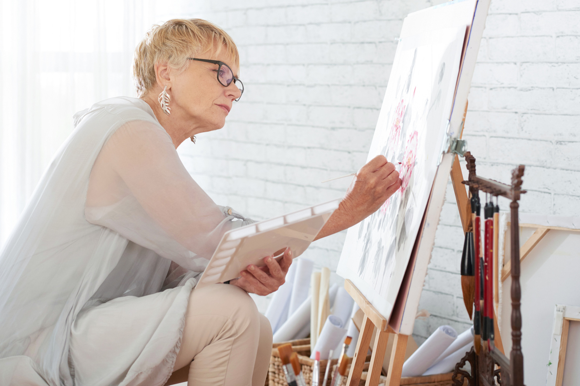 An older woman with short blonde hair and glasses is painting on a canvas at an easel. She is dressed in light-colored clothing, holding a palette, and appears focused on her artwork in a bright, airy studio.