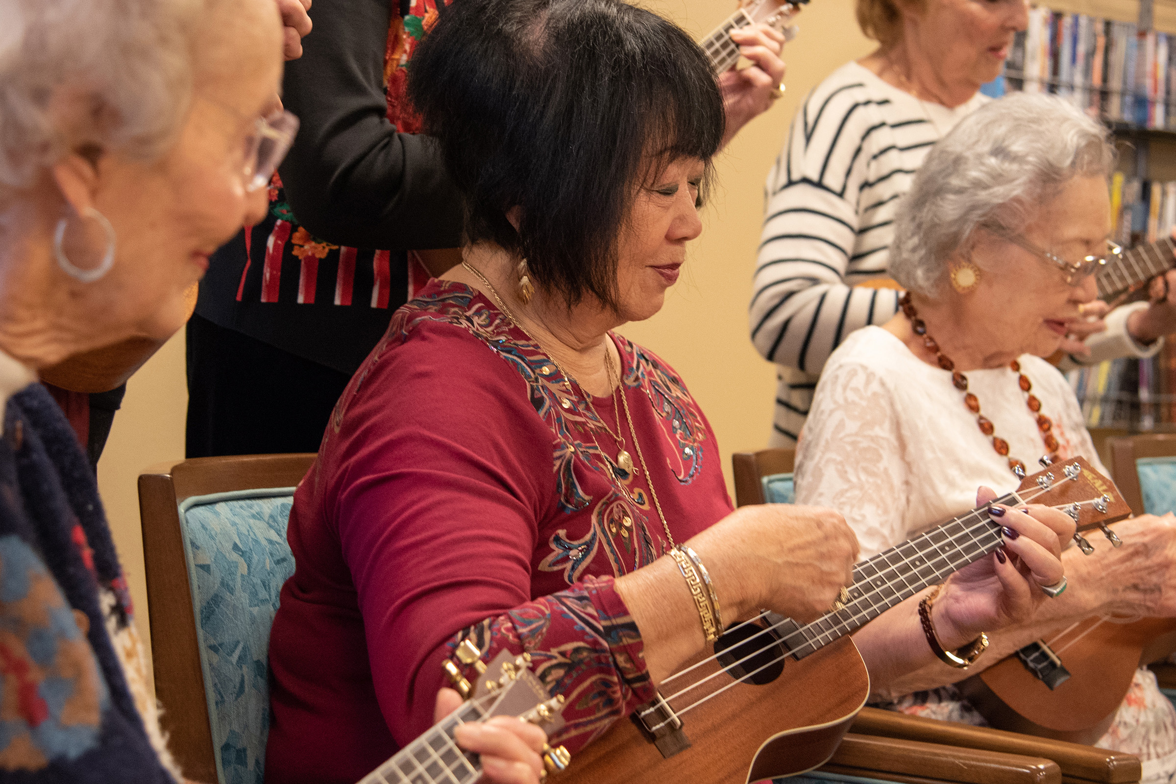 A group of elderly women sit in chairs playing ukuleles together, focused and smiling, in what appears to be a communal or recreational setting.