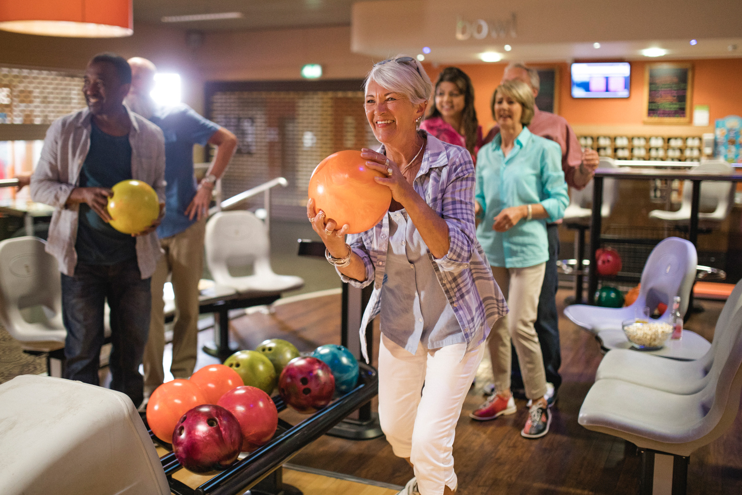 A smiling older woman prepares to bowl at a bowling alley, holding an orange ball while friends cheer behind her. Several colorful bowling balls are on the rack and the group appears happy and relaxed.