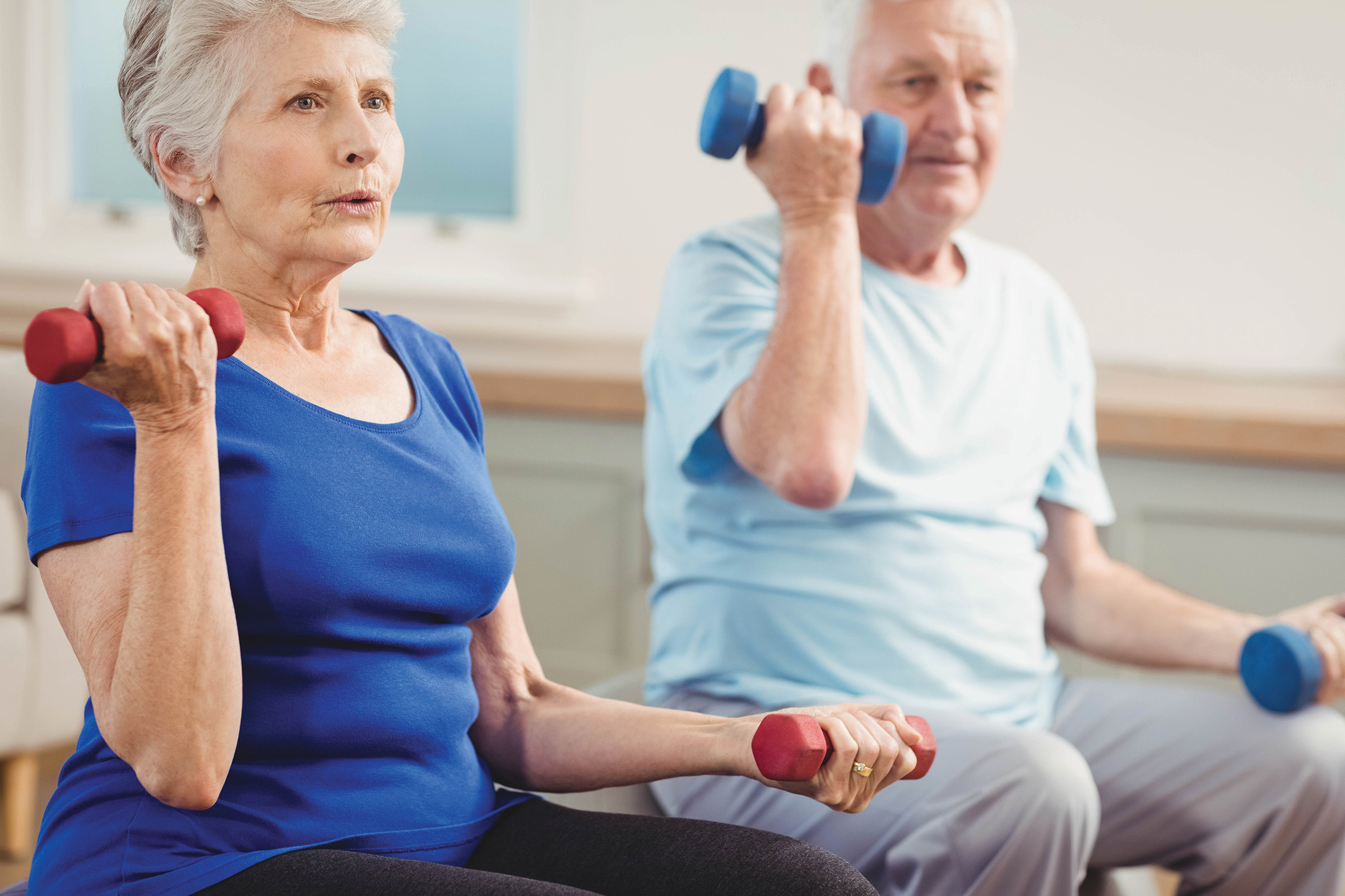 Two older adults sitting indoors, each holding a dumbbell in one hand and exercising. The woman in front wears a blue shirt and the man behind wears a light blue shirt. Both look focused on their workout.