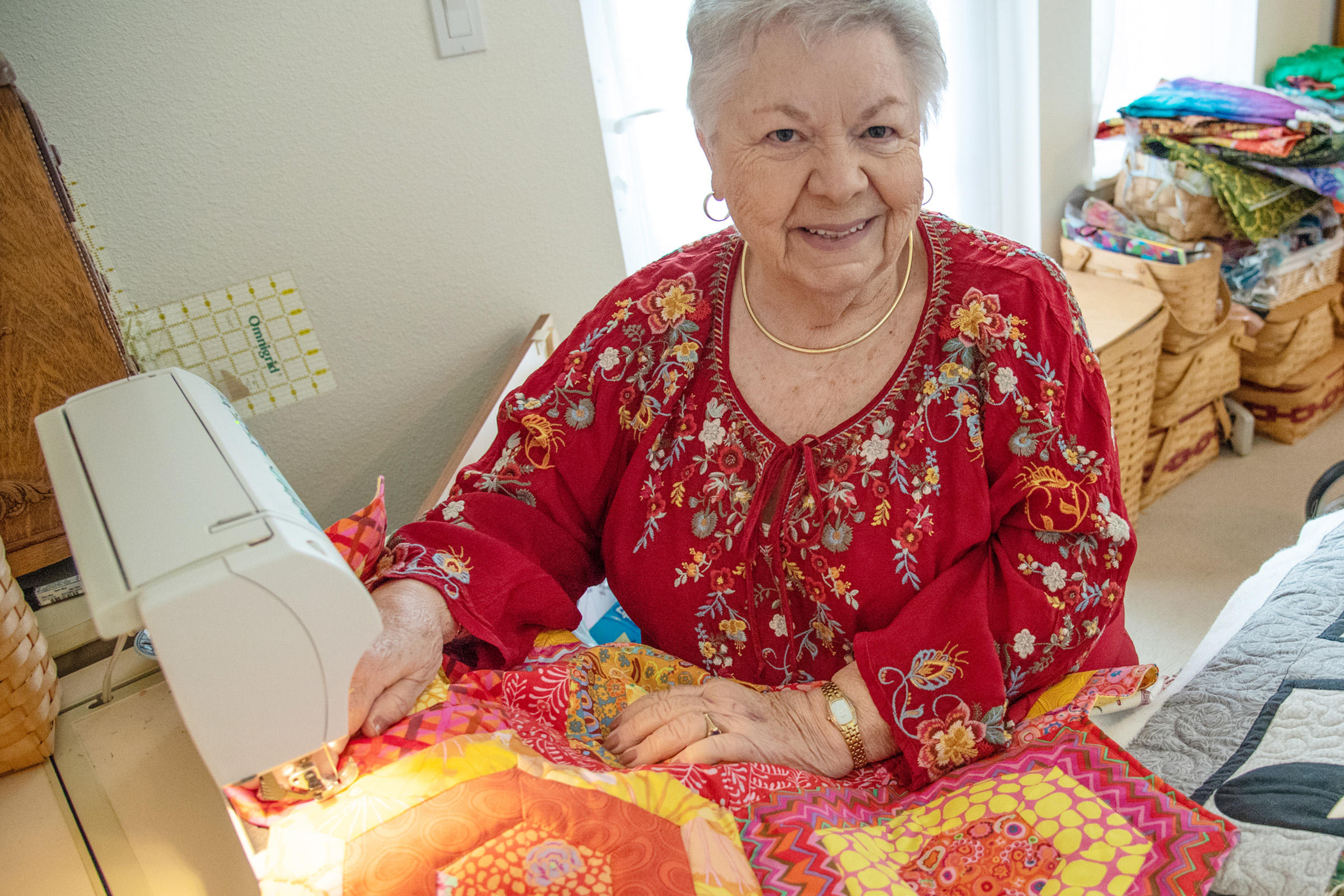 An older woman wearing a red, embroidered blouse smiles while sewing a colorful quilt at a sewing machine in a cozy, sunlit room with fabric and baskets in the background.
