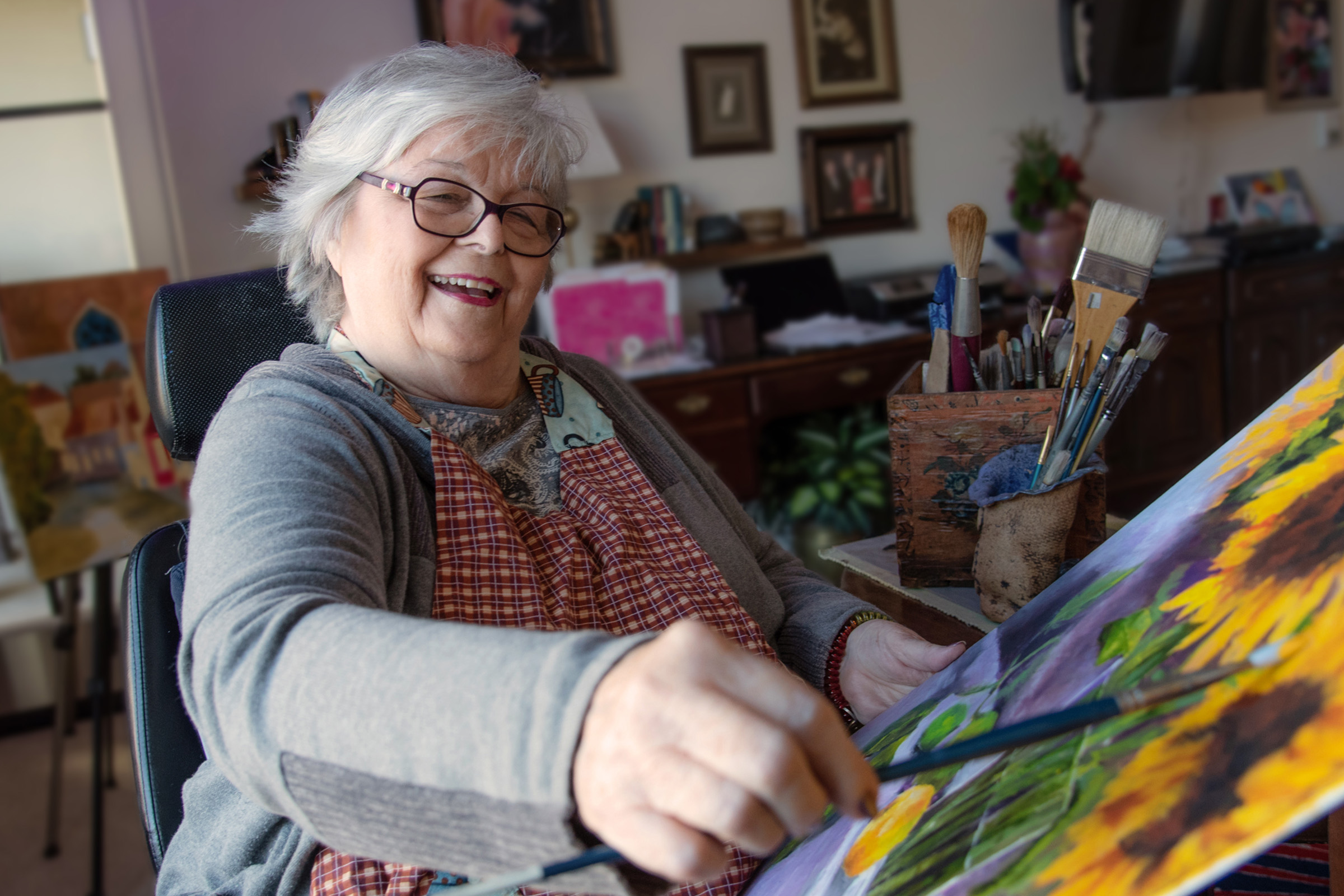 An older woman with gray hair and glasses smiles while painting a colorful canvas. She sits indoors at a desk with brushes and art supplies, wearing an apron. Her surroundings include framed pictures and a cozy, homey atmosphere.
