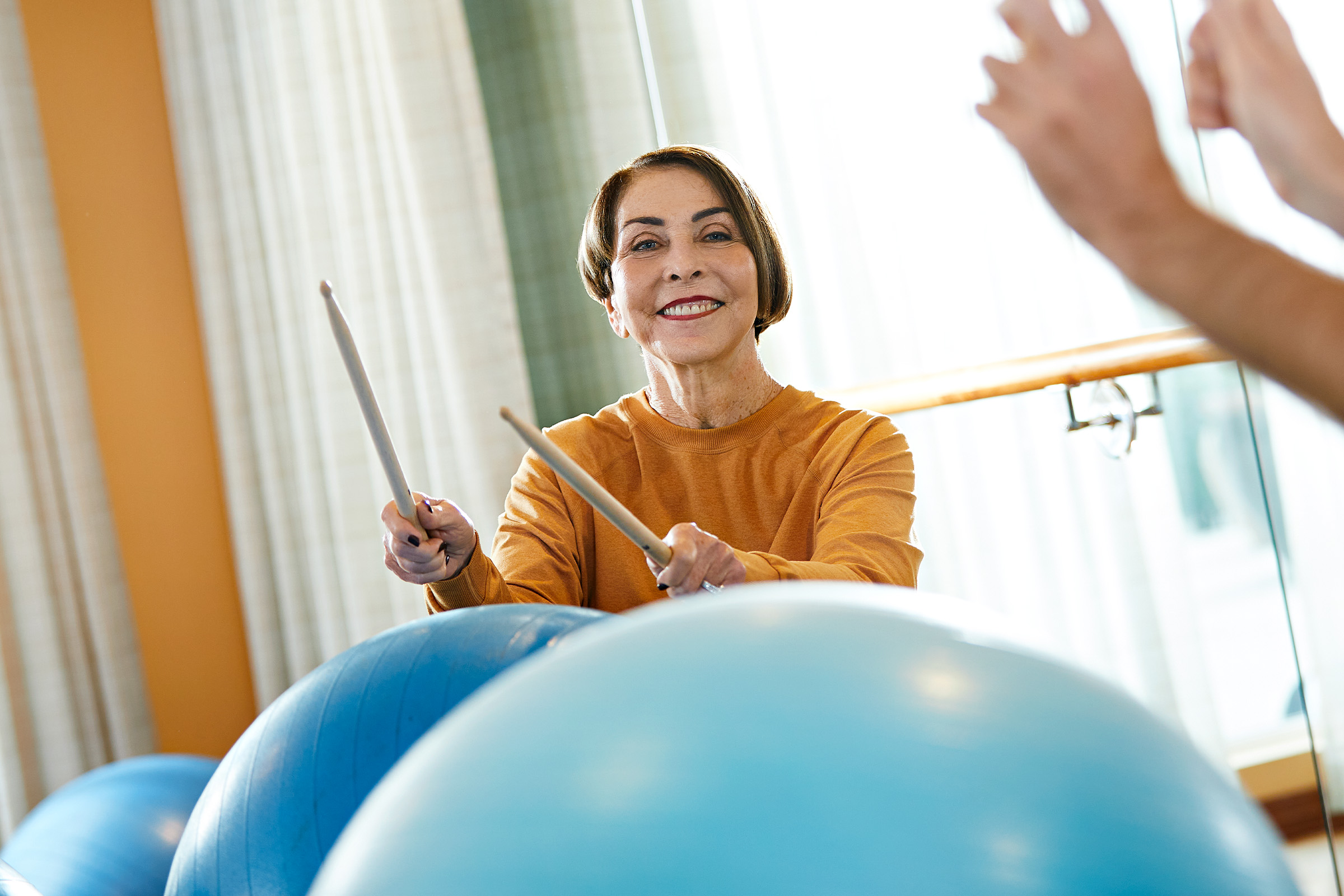 A smiling woman in an orange top holds drumsticks and plays on large blue exercise balls in a bright room, participating in a fitness or drumming exercise class.