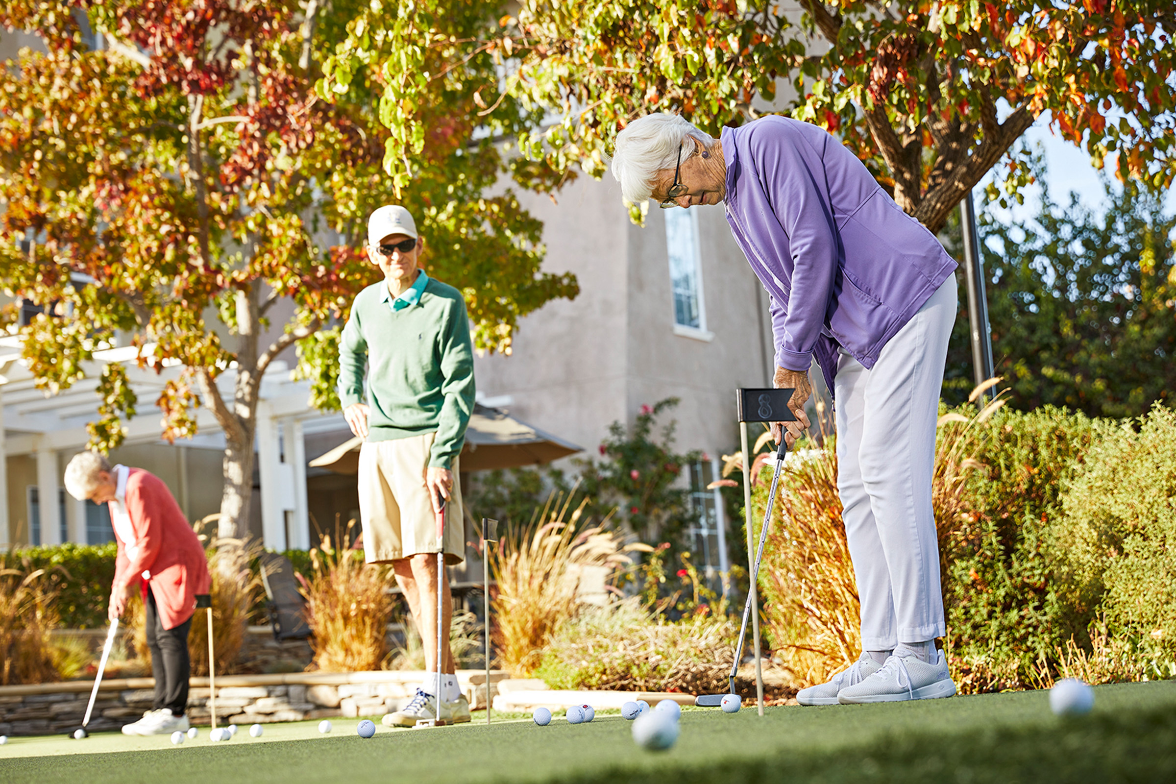 Three older adults are playing mini-golf on a sunny day. One woman in a purple jacket is putting, while another woman and a man in casual clothes watch and play nearby. Trees with fall foliage are in the background.