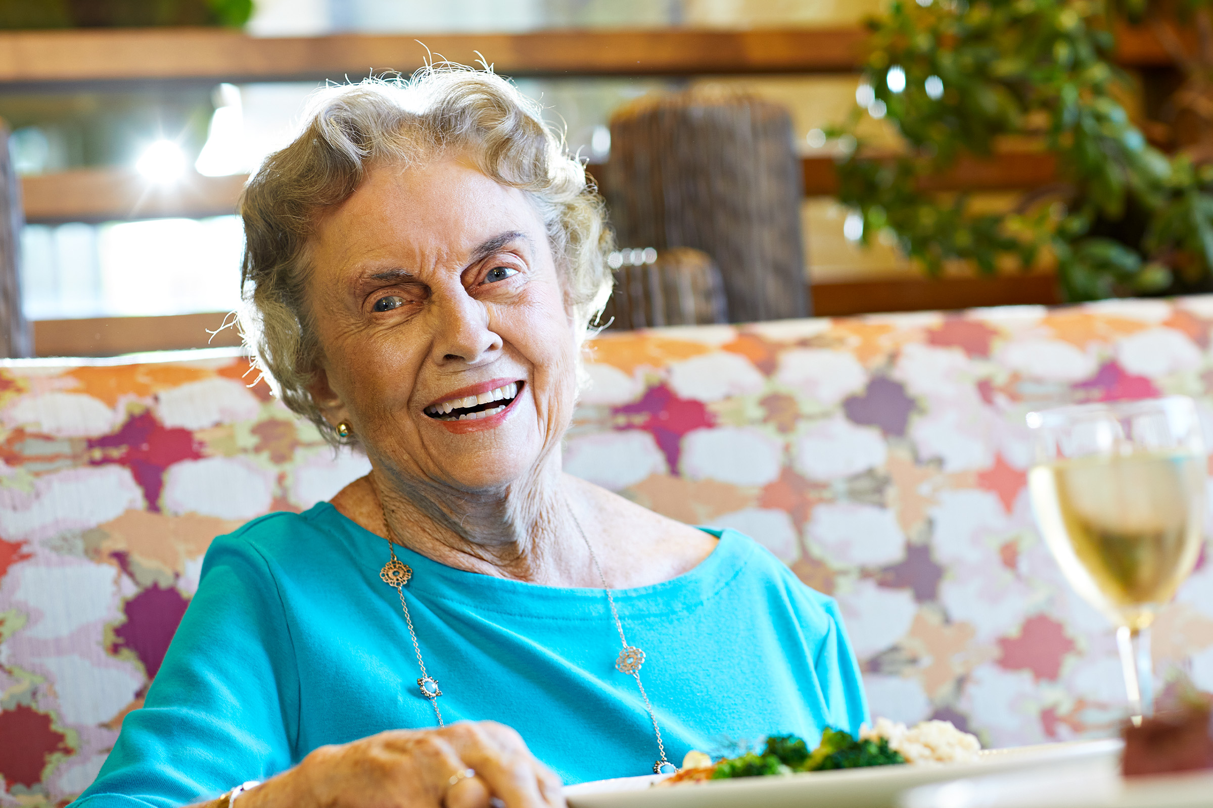 An elderly woman with short gray hair and wearing a turquoise top sits smiling at a table with food and a glass of white wine in front of her. The background is bright and colorful.