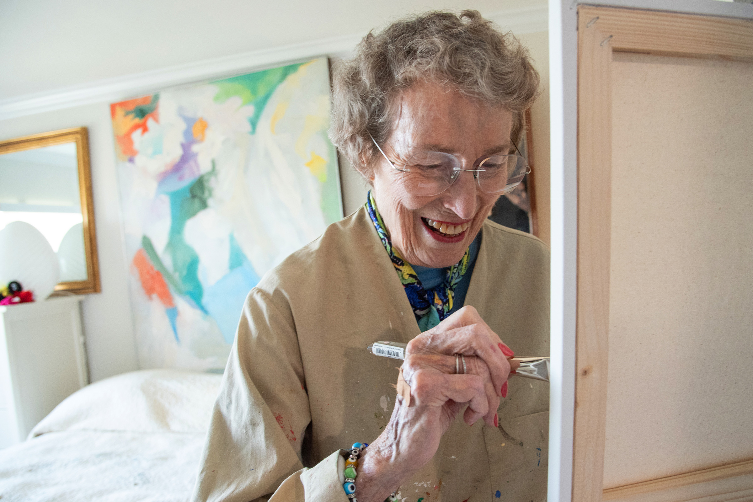 An elderly woman wearing glasses and a paint-splattered smock smiles while painting on a canvas. A colorful abstract painting hangs on the wall behind her in a bright, cozy room.
