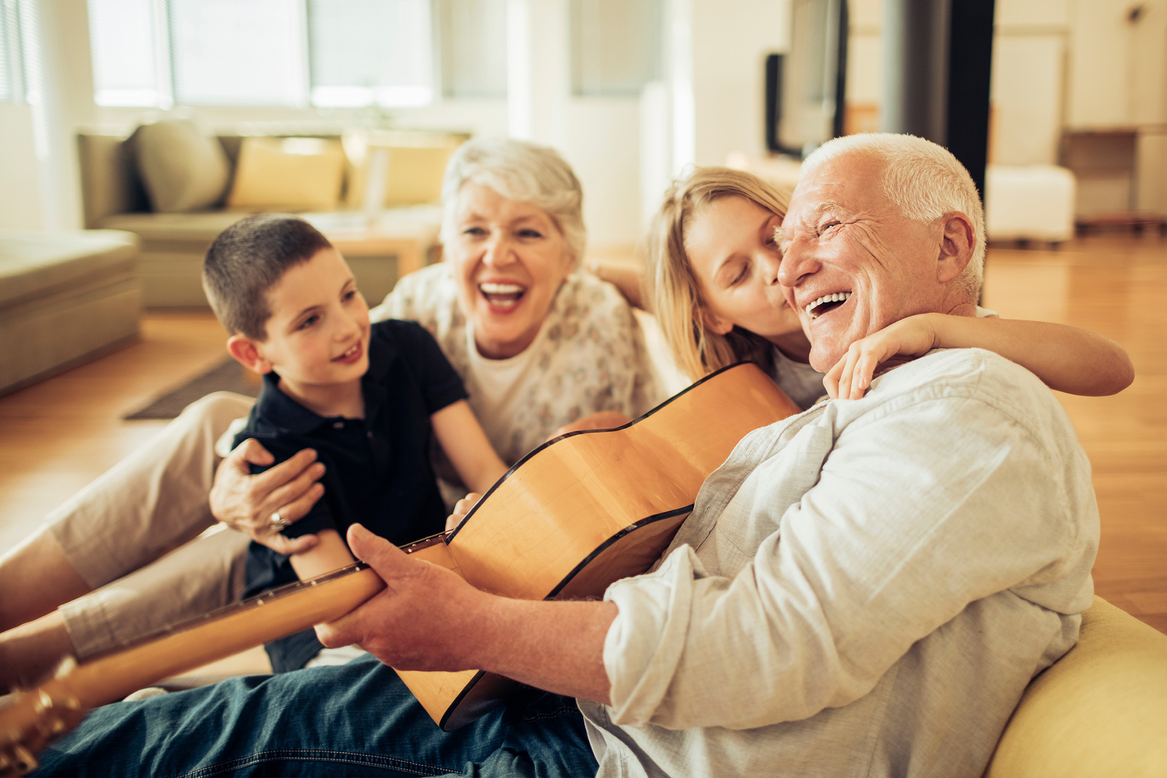A joyful elderly man plays guitar while sitting on the floor with a smiling elderly woman and two children; the girl hugs and kisses him, while the boy sits nearby. Sunlight fills the cozy living room.