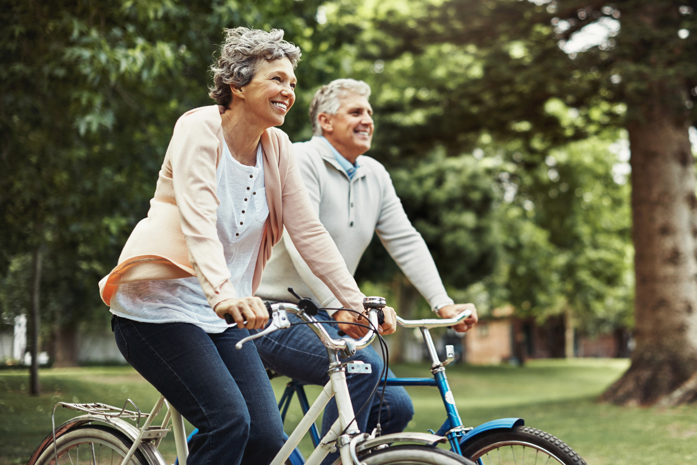 Two smiling older adults ride bicycles together in a park, surrounded by green trees. They appear relaxed and happy, enjoying the outdoors on a bright day.