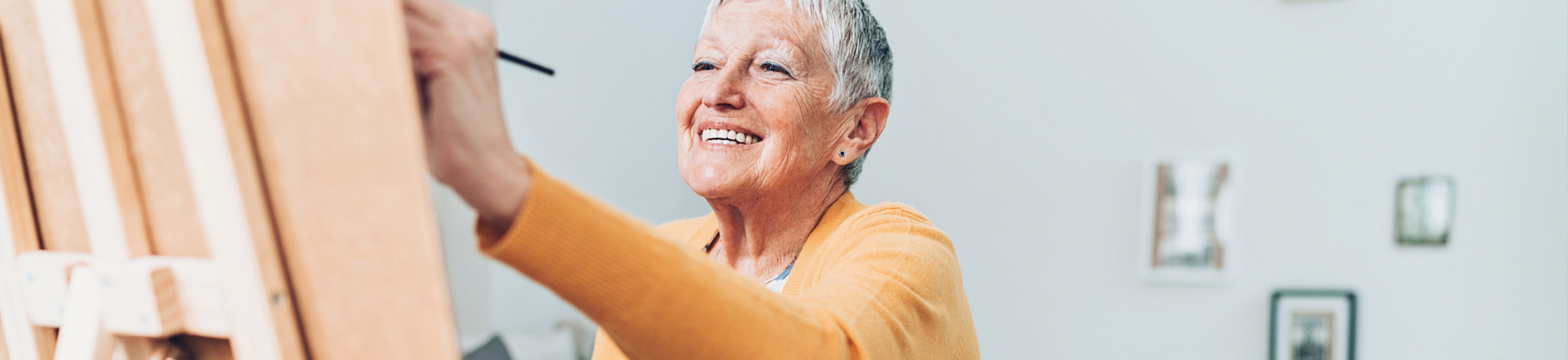 An older woman with short gray hair, wearing a yellow cardigan, smiles while painting at an easel in a bright room.
