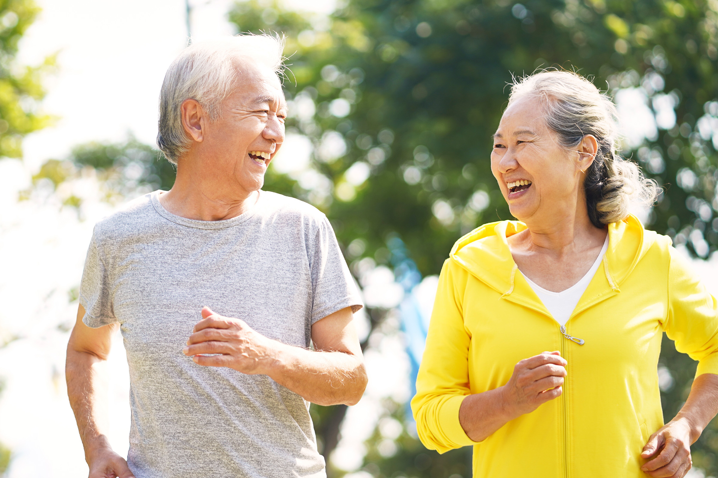 Two older adults, a man in a gray t-shirt and a woman in a yellow jacket, are jogging outdoors together, smiling and looking at each other, with trees and sunlight in the background.