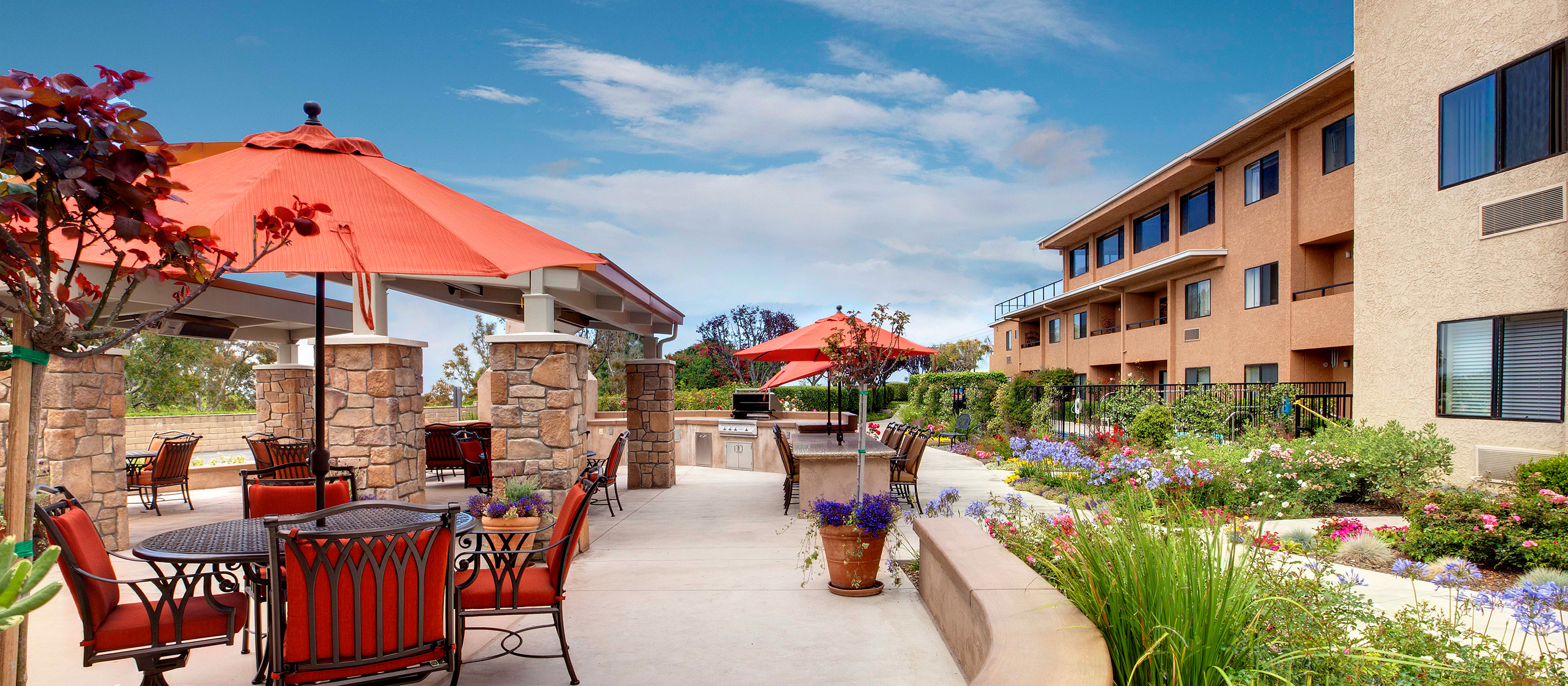 Outdoor patio area with tables, chairs, and red umbrellas, surrounded by stone pillars and colorful flower beds, next to a tan-colored apartment building under a blue sky with clouds.