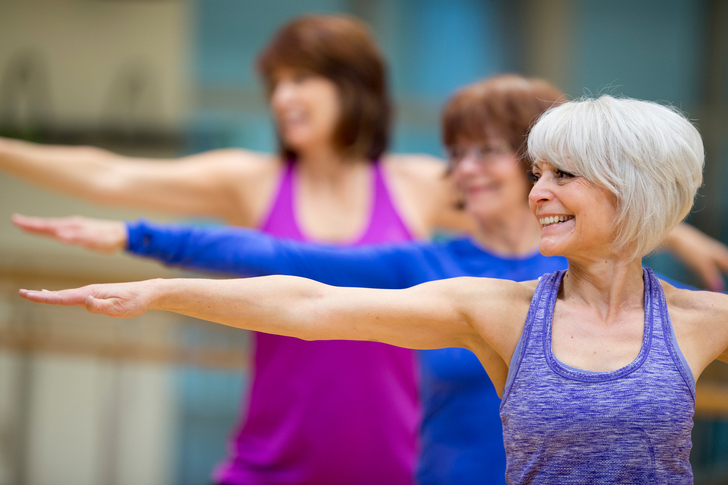 Three women in fitness attire practice a yoga pose indoors, standing in a row with arms extended and smiling. The focus is on a woman with short gray hair in the foreground.