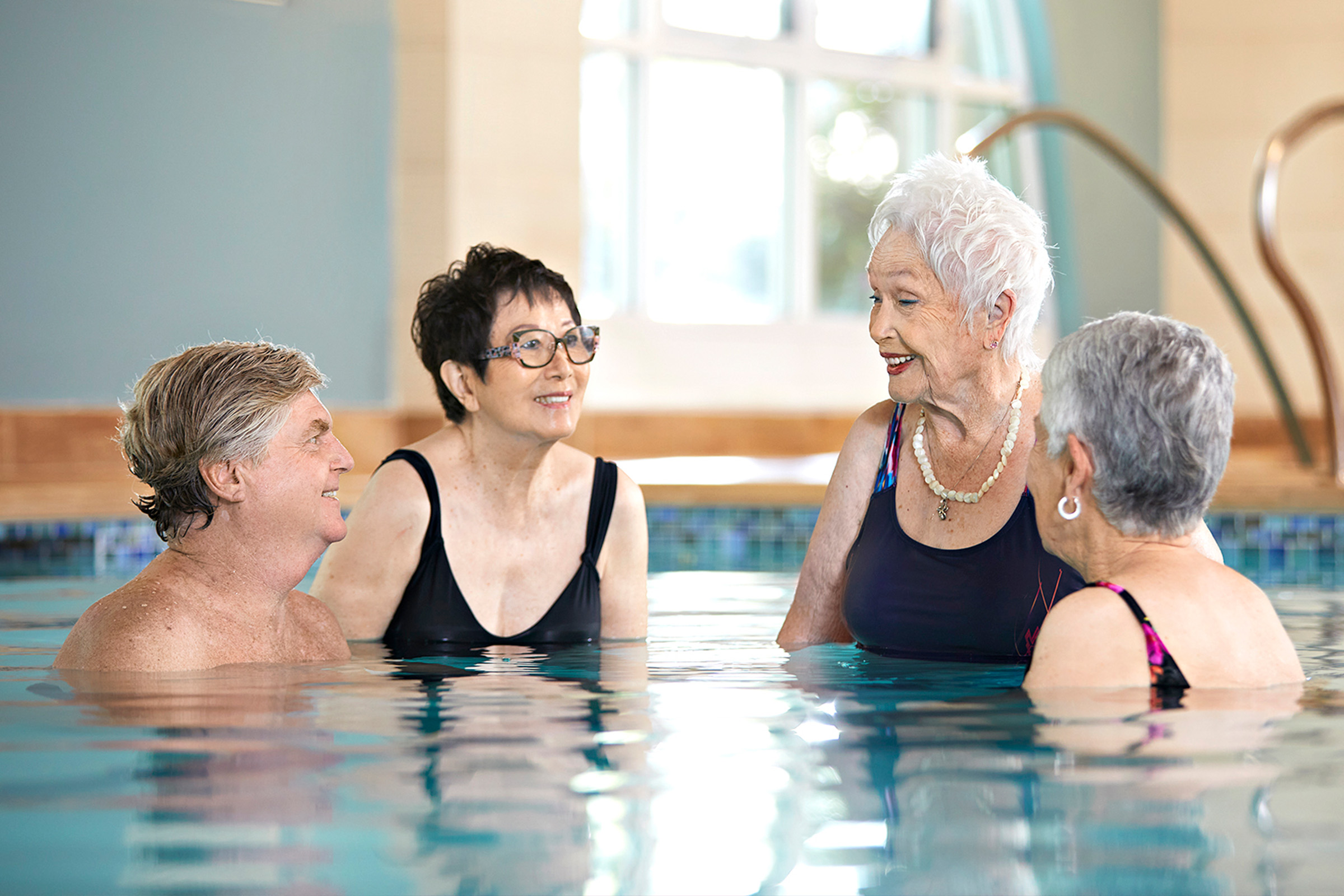 Four older adults are standing and talking together in an indoor swimming pool, smiling and enjoying each others company near the pools edge with stairs visible in the background.